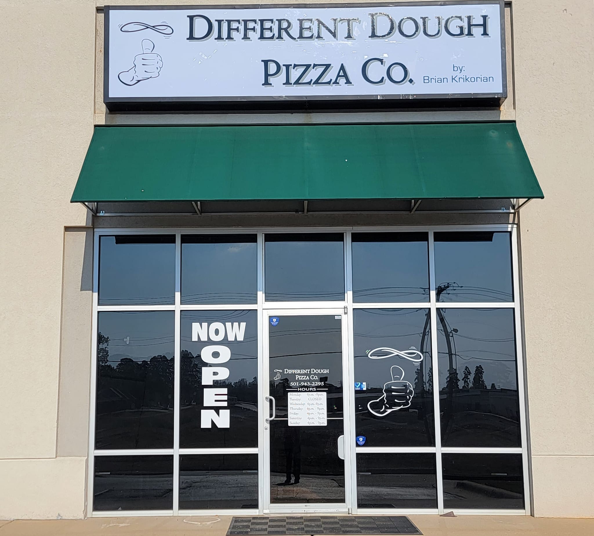 2 - Different Dough Pizza Co Storefront of “Different Dough Pizza Co.” with a green awning, large glass doors, and “NOW OPEN” signs displayed. The logo features a hand holding pizza dough. The area outside is sunny with a clear view of the street.