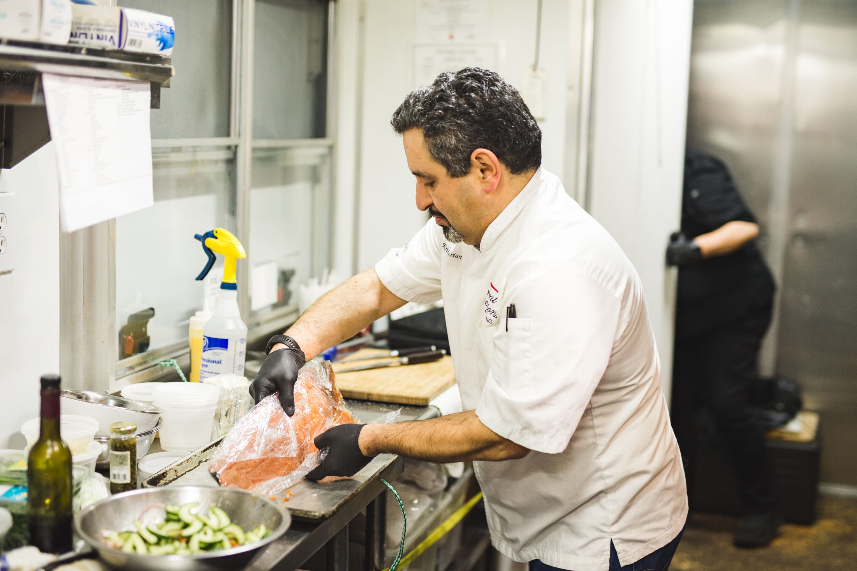 Little Rock Arkansas Wedding Photographer A chef wearing a white coat and black gloves prepares a large piece of salmon in a commercial kitchen, while another person works in the background. There are ingredients and utensils on the counter.