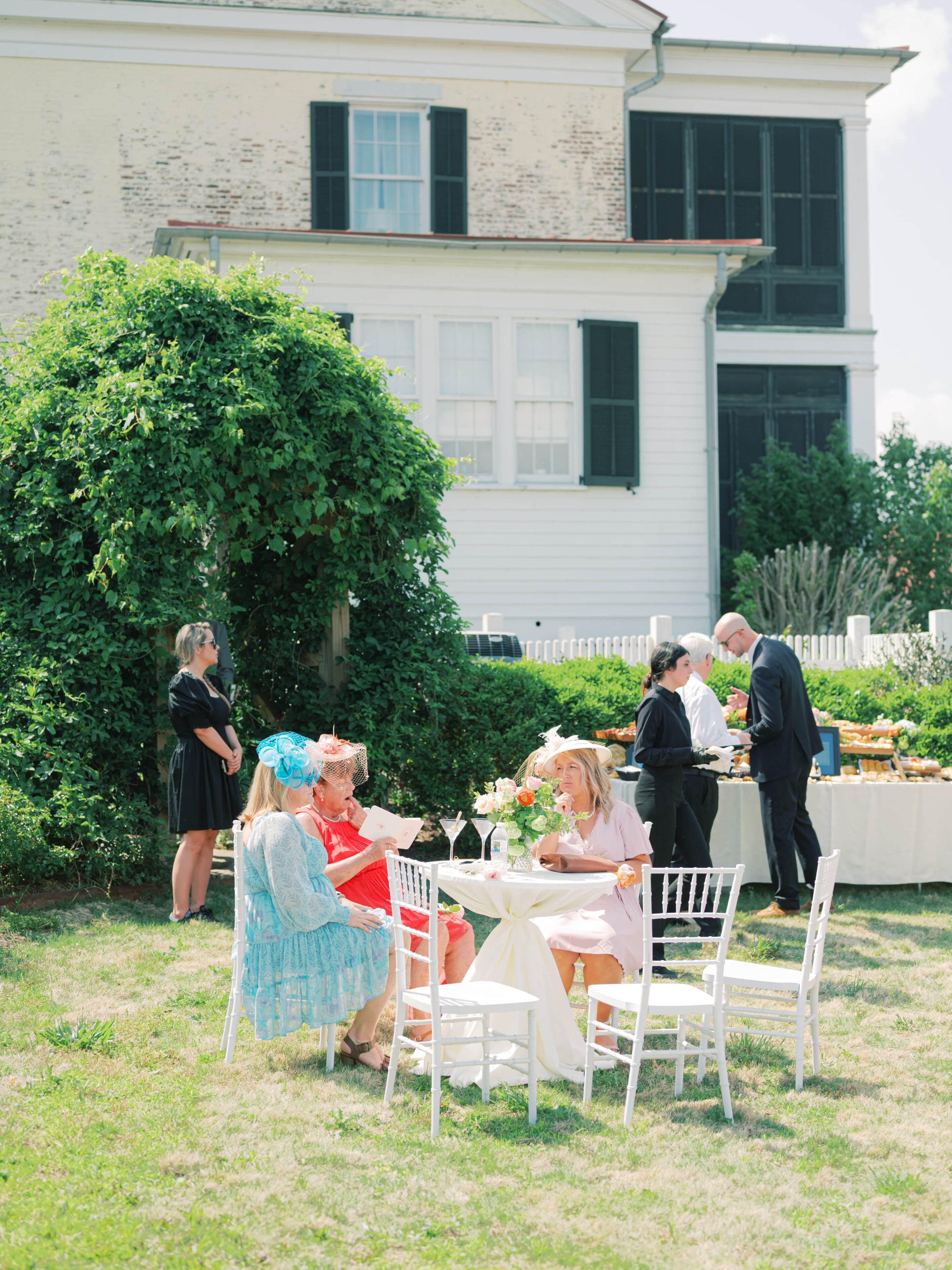Liz and Matt Wedding Three women in colorful dresses and hats sit at a round table outdoors, chatting and eating. Two servers in black stand nearby, with a buffet setup behind them. A large house and greenery are in the background.