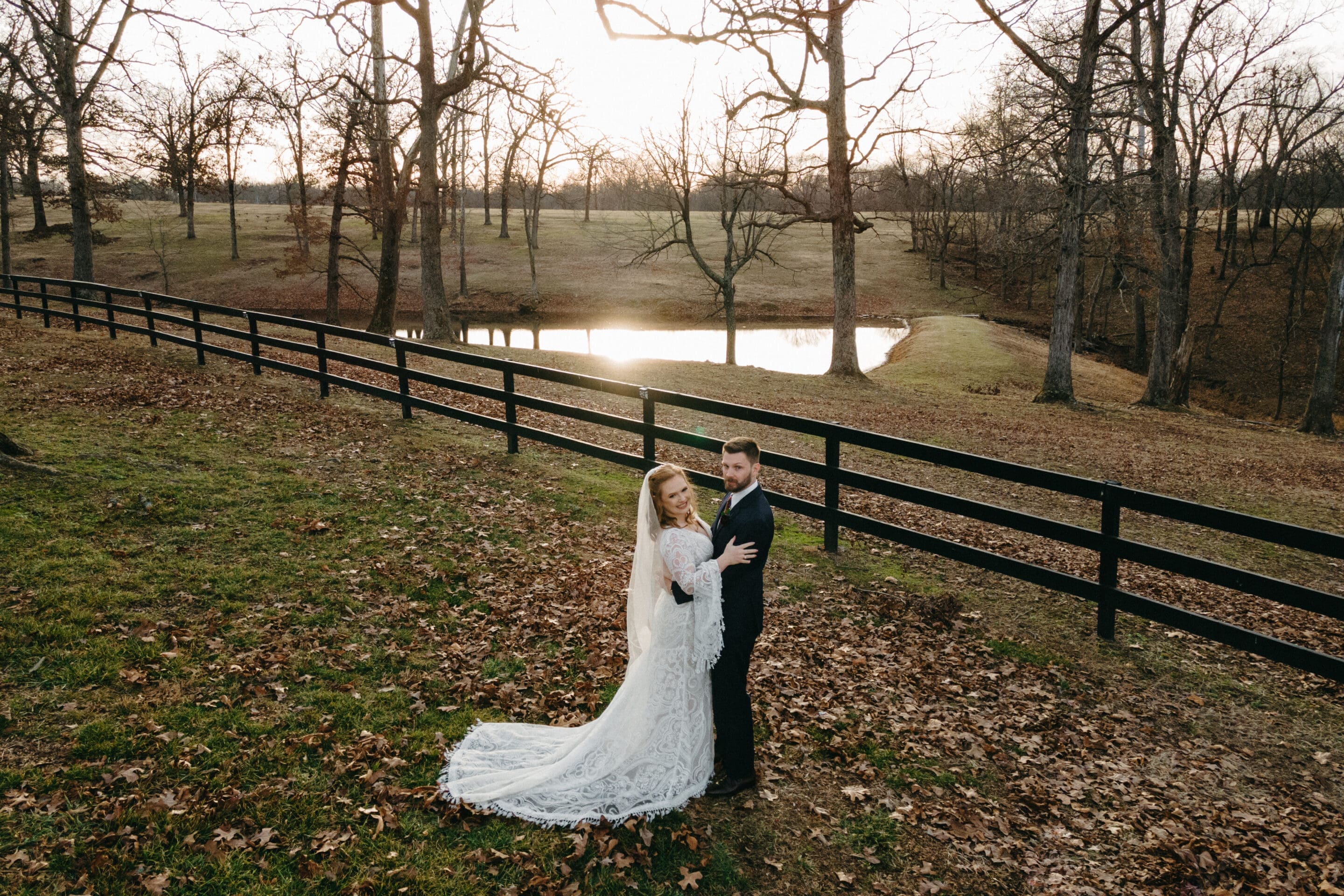 John David + Lindsey A bride and groom in wedding attire stand together on a grassy hill covered with autumn leaves, near a black wooden fence and a small pond, with bare trees and a sunset in the background.