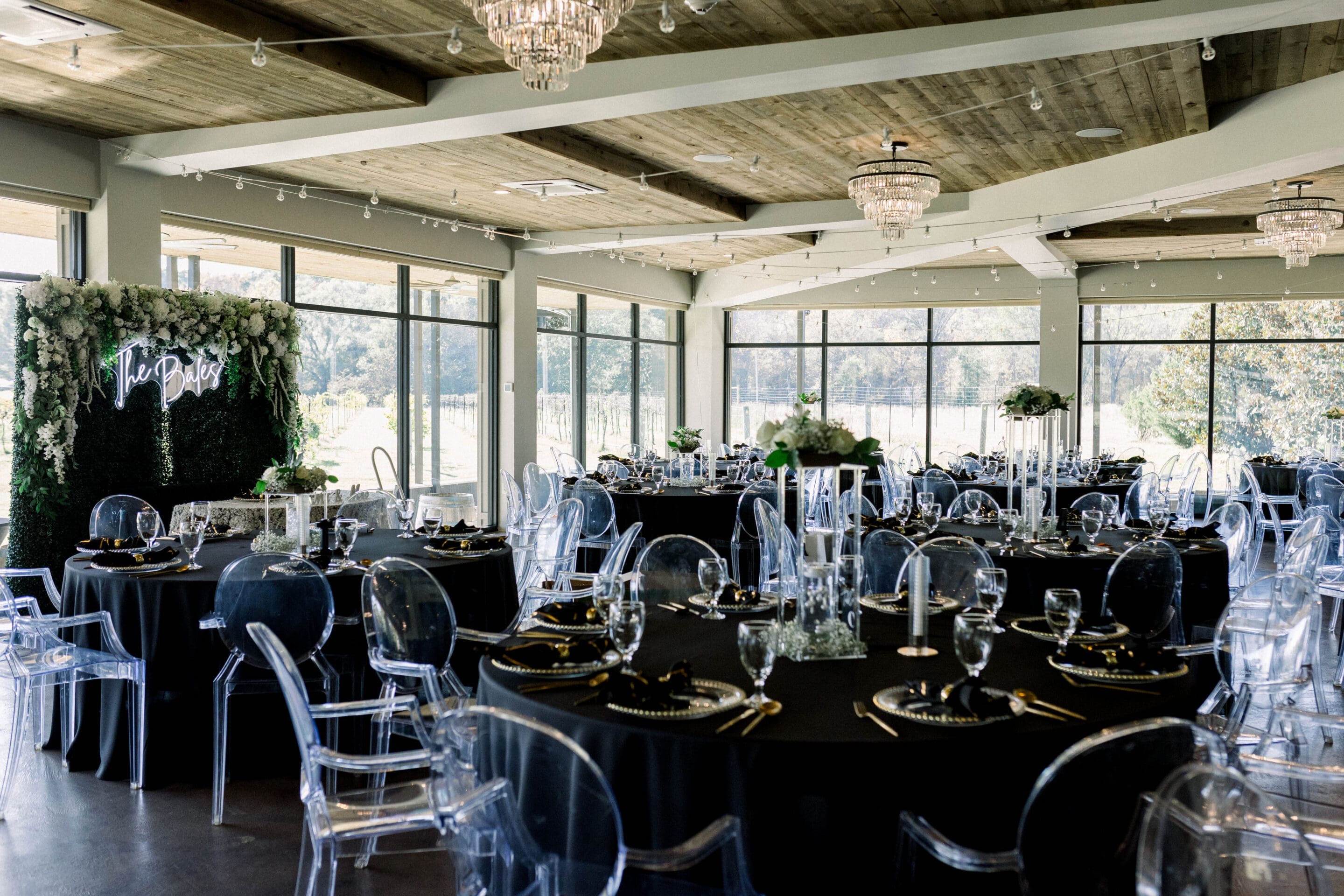 Keely + Joshua Elegant event venue with round tables covered in black tablecloths, clear acrylic chairs, floral arrangements, glassware, and a flower-covered backdrop with a neon sign. Large windows let in natural light.