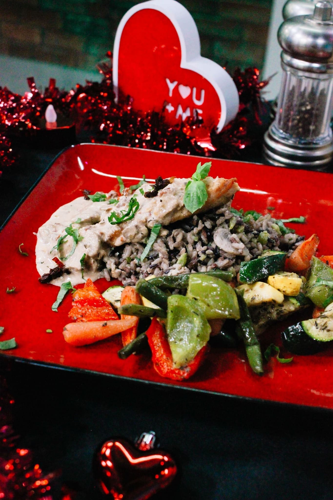 2 - Valentine_s Day Cook At Home A festive meal on a red plate features chicken in creamy sauce over rice with sautéed carrots, zucchini, and peppers. Valentine’s Day decor, red tinsel, and a heart with YOU + ME are in the background.