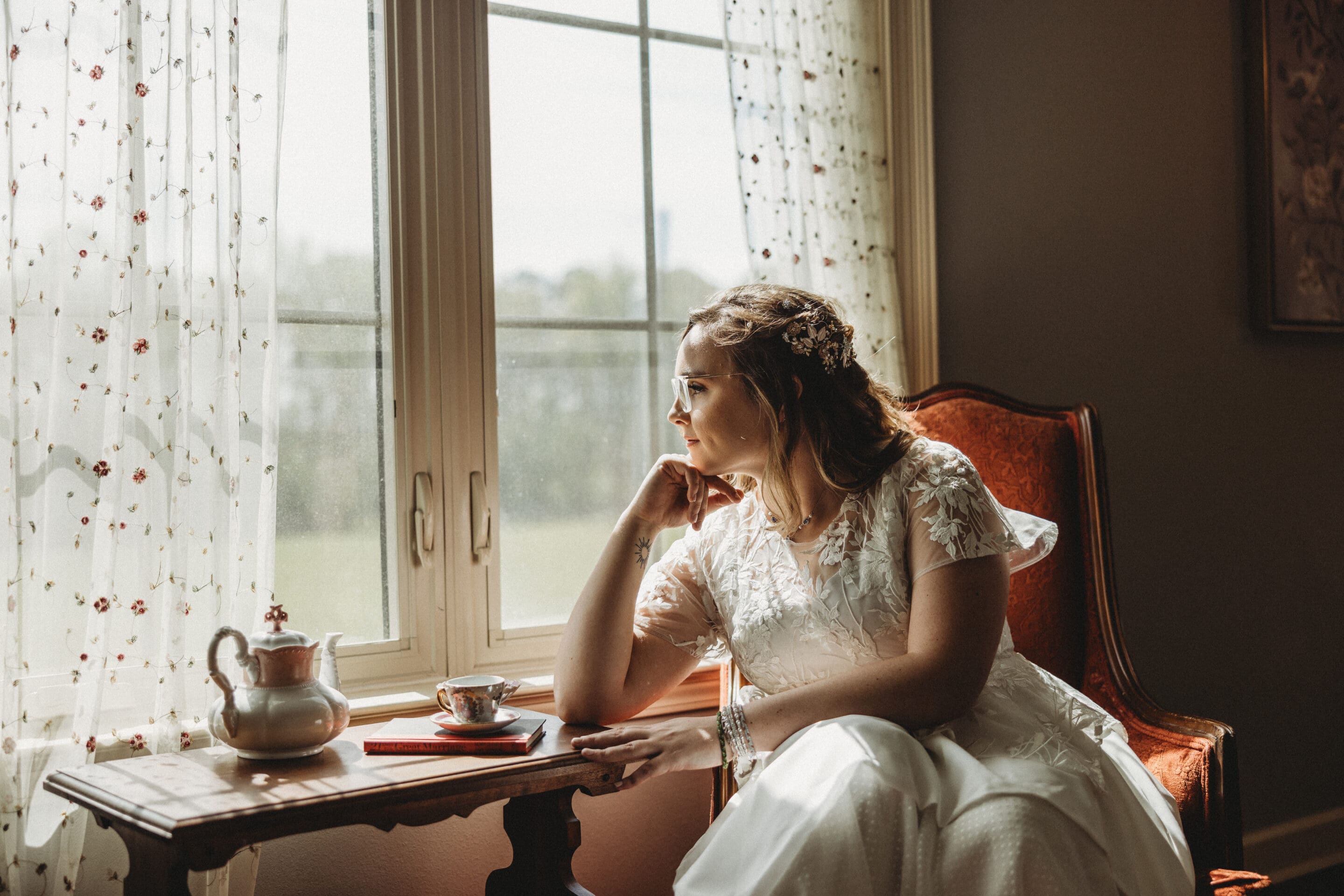A woman in a white dress sits in an armchair by a window, gazing outside thoughtfully. A teapot and teacup rest on a small wooden table beside her. Sunlight filters through lace curtains.
