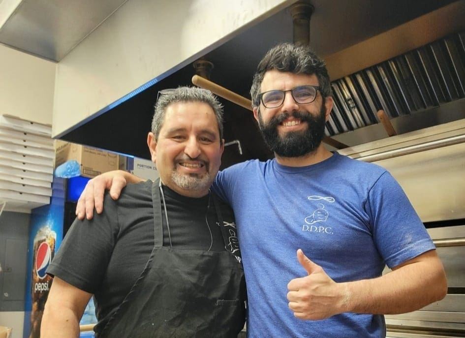 3 - Chef Serge Krikorian Brian Krikorian Two men stand together in a kitchen, smiling at the camera. One wears a black apron and the other, in a blue shirt, gives a thumbs up. Stainless steel kitchen equipment and a Pepsi machine are visible in the background.