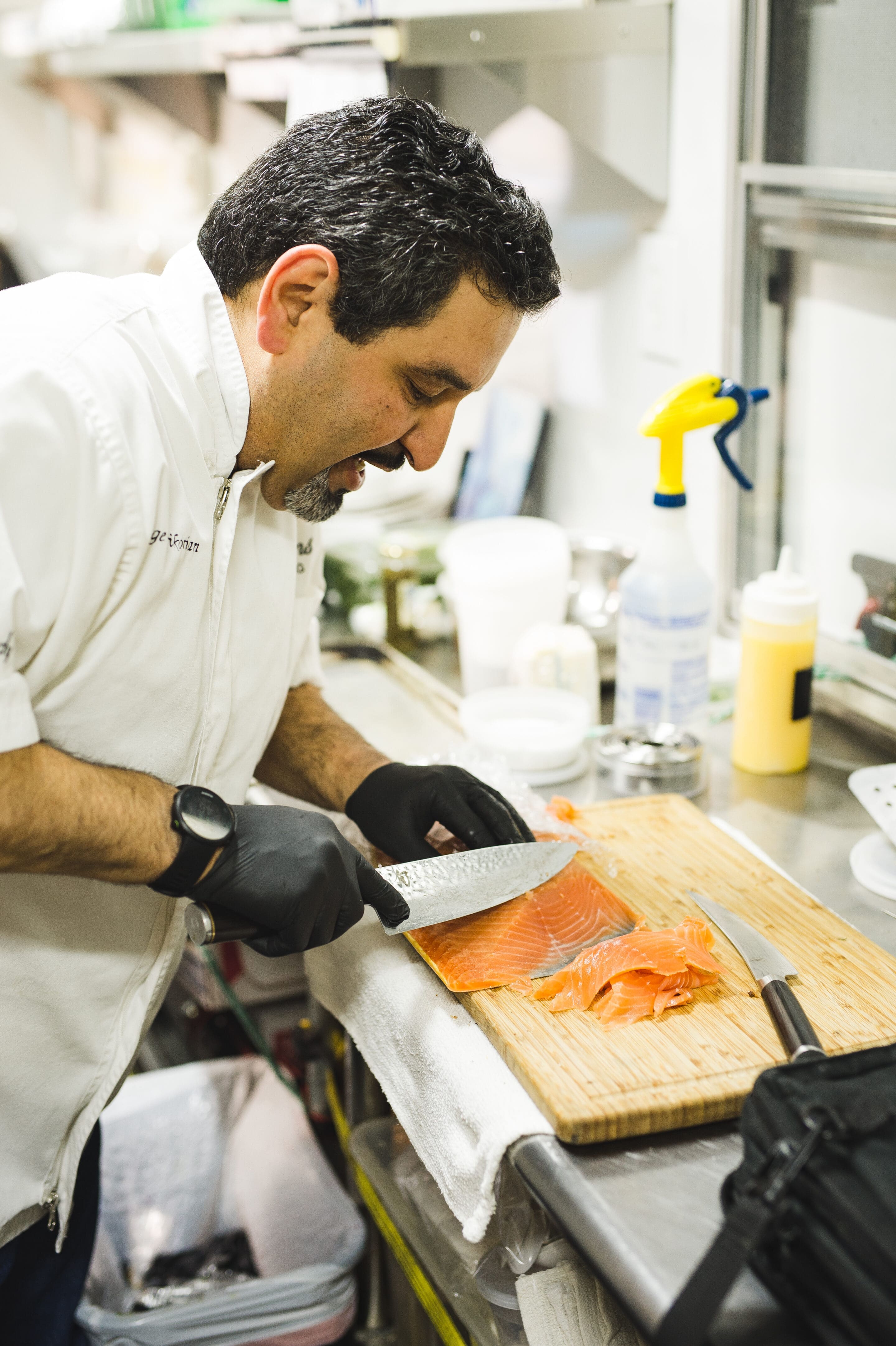 Little Rock Arkansas Wedding Photographer A chef wearing a white coat and black gloves slices a fillet of salmon on a wooden cutting board in a commercial kitchen. Various kitchen tools and containers are visible in the background.