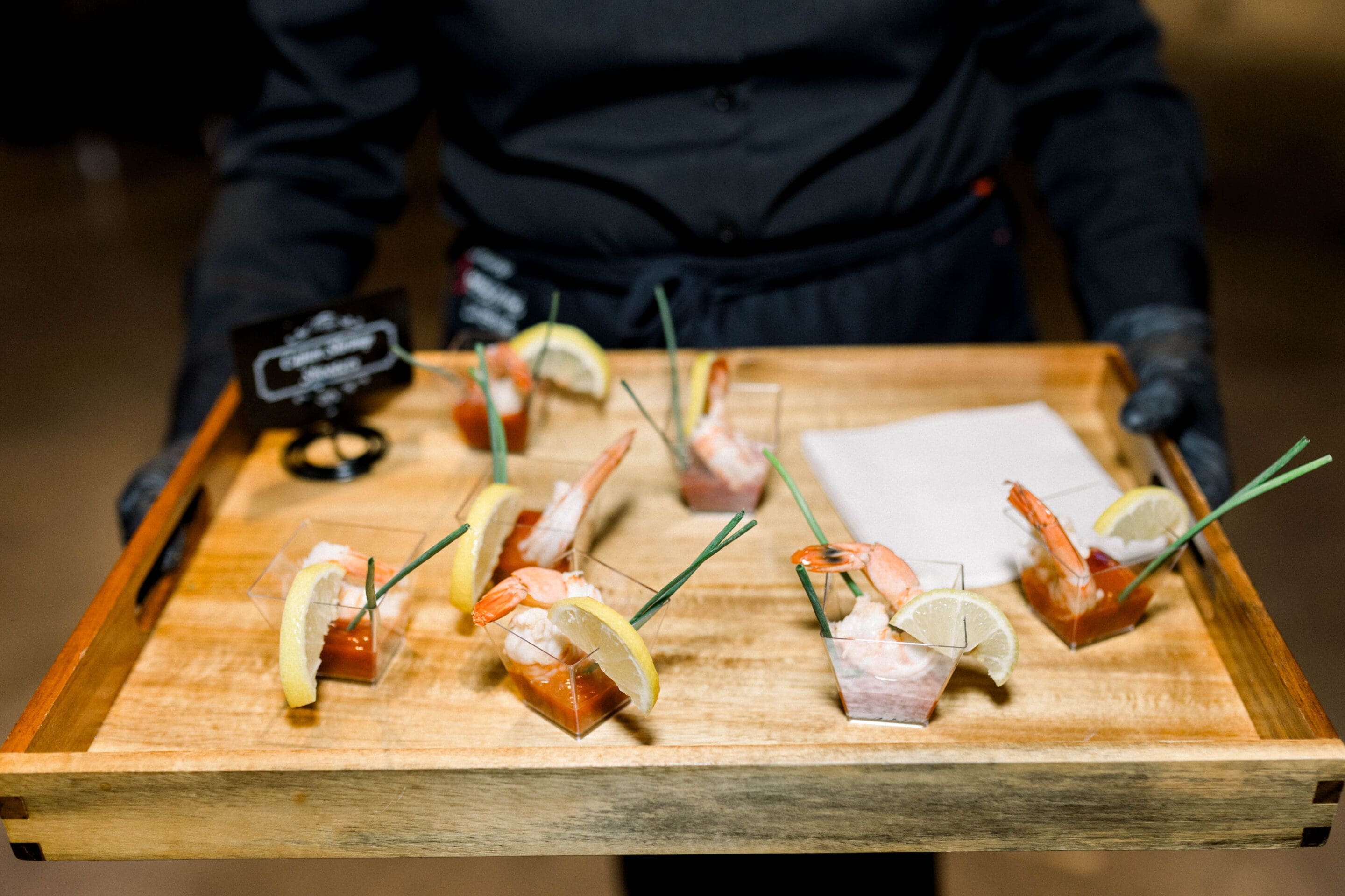Keely + Joshua A server in black attire holds a wooden tray with cocktail shrimp in small clear cups, garnished with lemon slices and green chives, alongside cocktail sauce and neatly folded napkins.