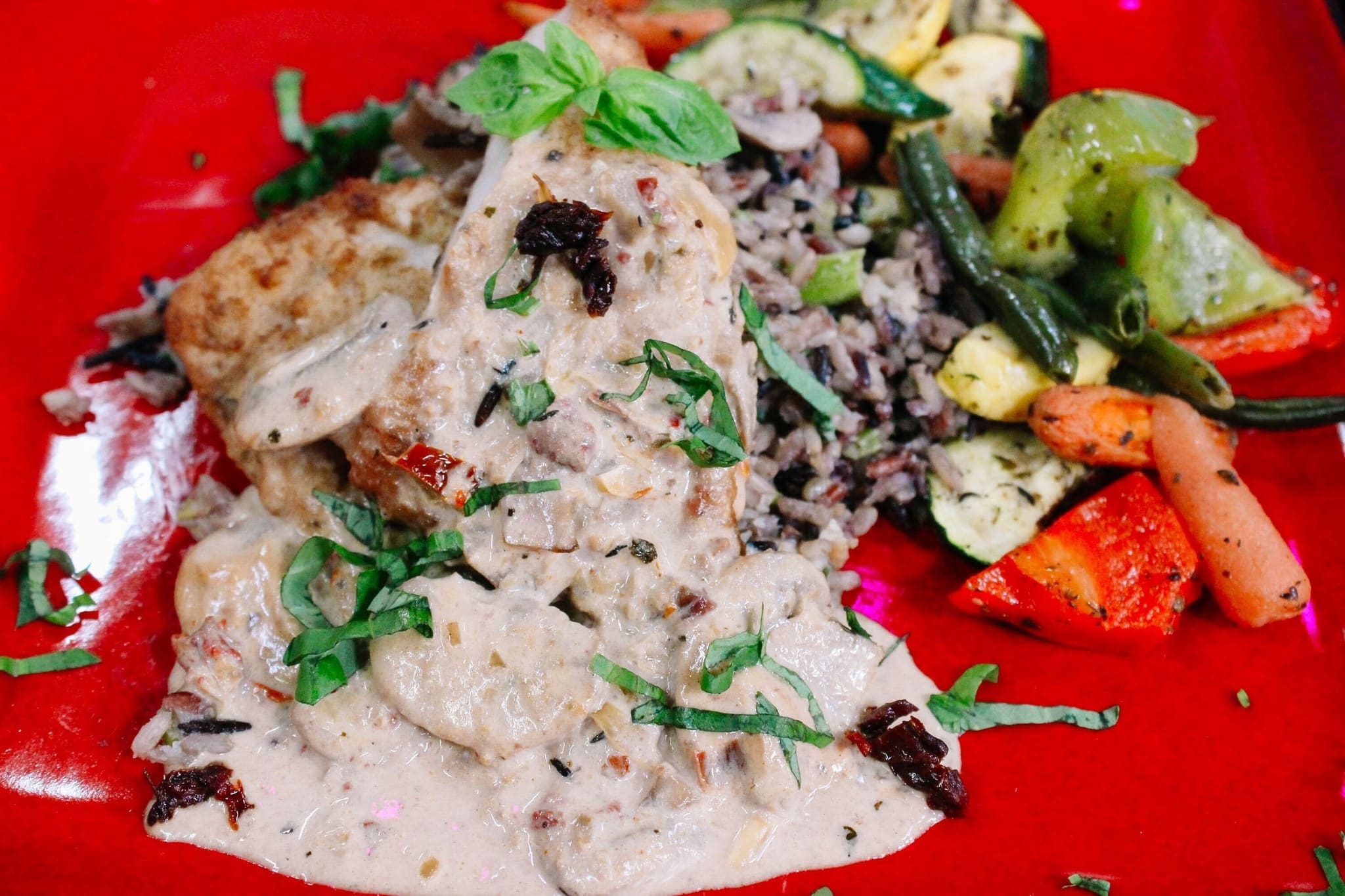 3 - Marry Me Chicken Recipe A plate of creamy chicken with herbs, served over wild rice with sautéed mixed vegetables including carrots, zucchini, and green beans, garnished with fresh basil on a red plate.