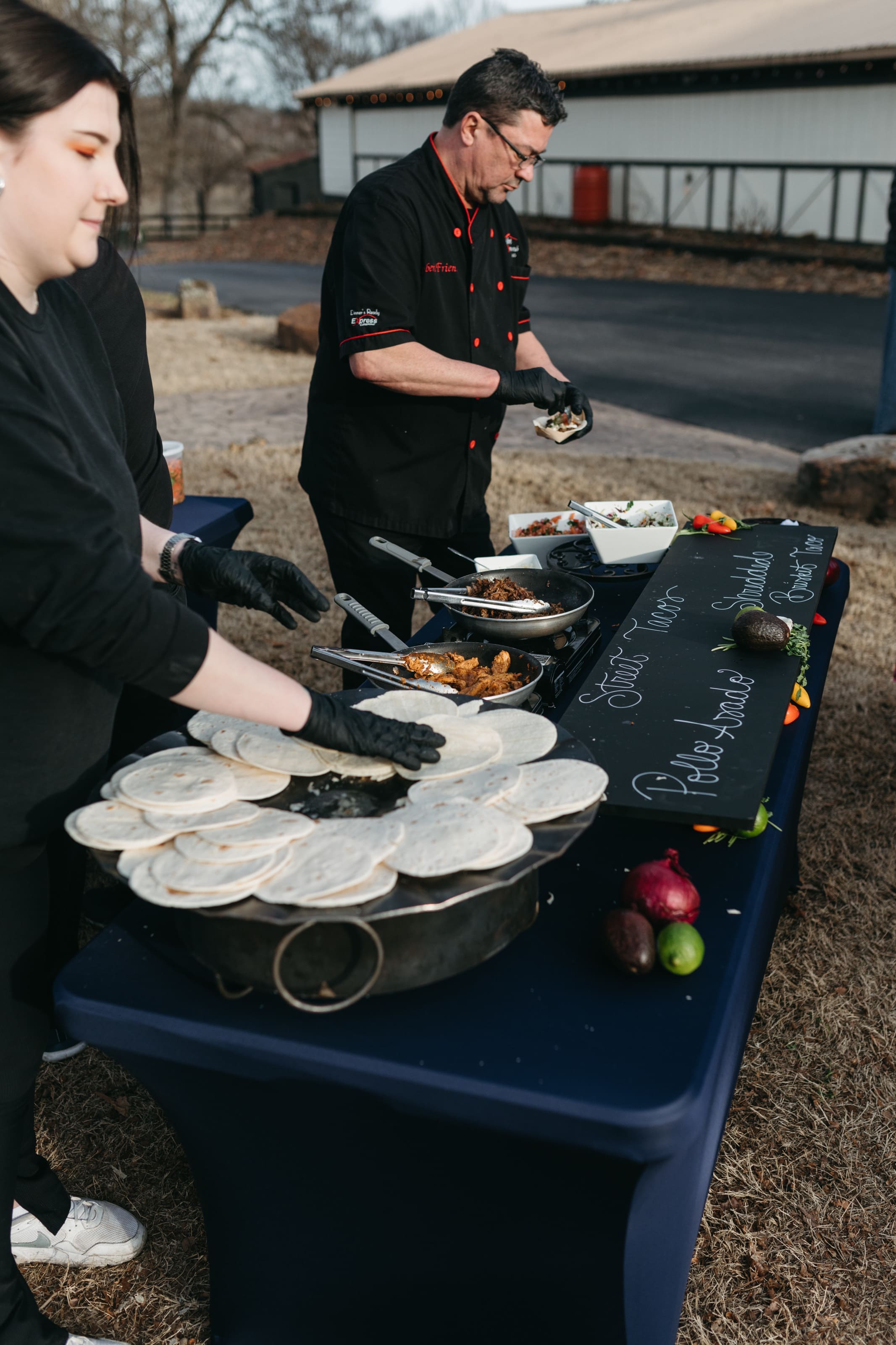 John David + Lindsey Two people prepare tacos at an outdoor food station with stacks of tortillas, various fillings, and a chalkboard menu. Both wear black clothing and gloves, and the scene is set on a grassy area near a building.