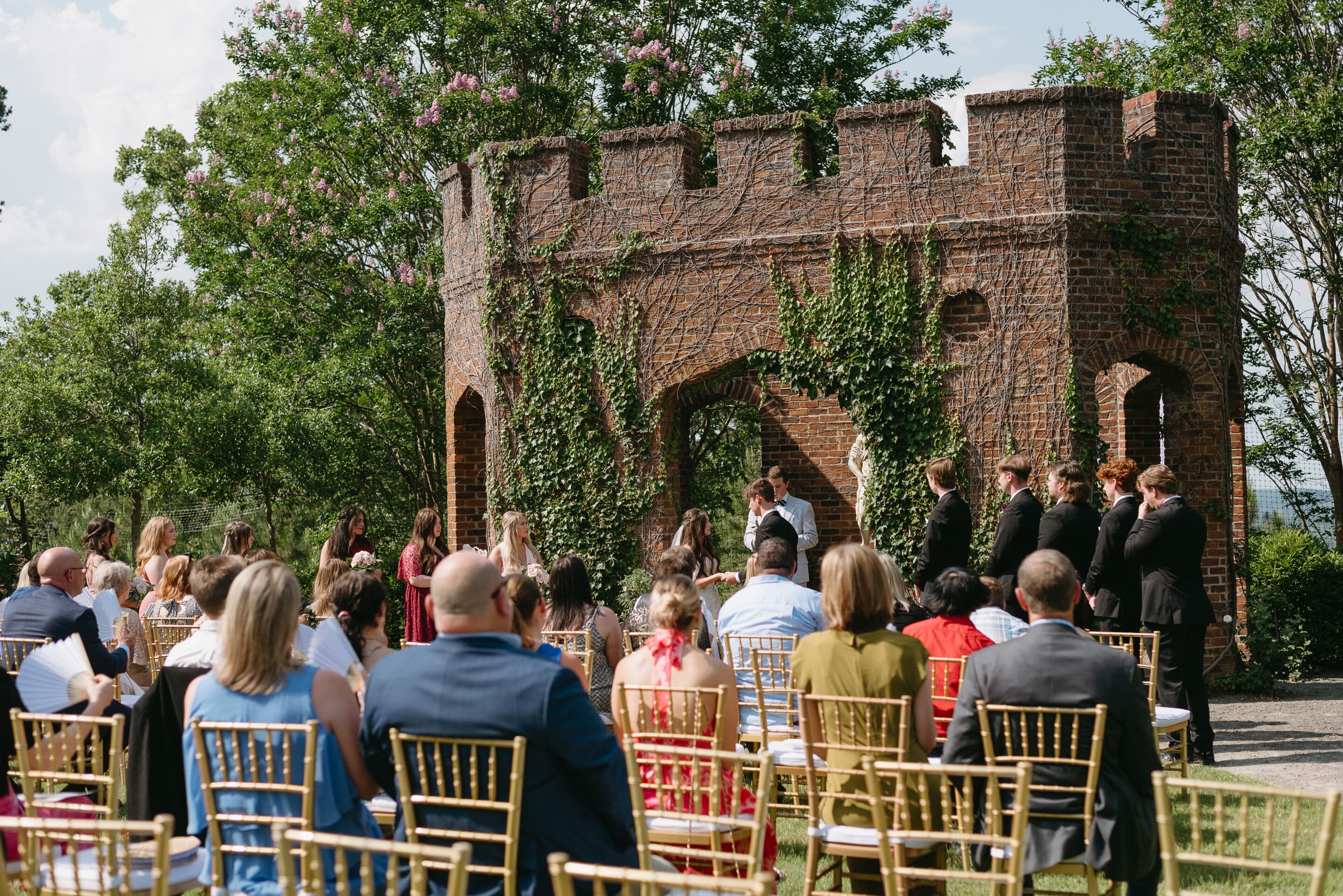 Lauren & Christian An outdoor wedding ceremony with guests seated on gold chairs facing a couple and officiant in front of a brick, ivy-covered castle-like structure, surrounded by greenery on a sunny day.