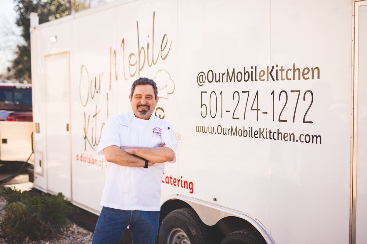Little Rock Arkansas Wedding Photographer A man wearing a white chefs jacket stands smiling with arms crossed in front of a white food truck labeled Our Mobile Kitchen with contact information and website printed on the side.