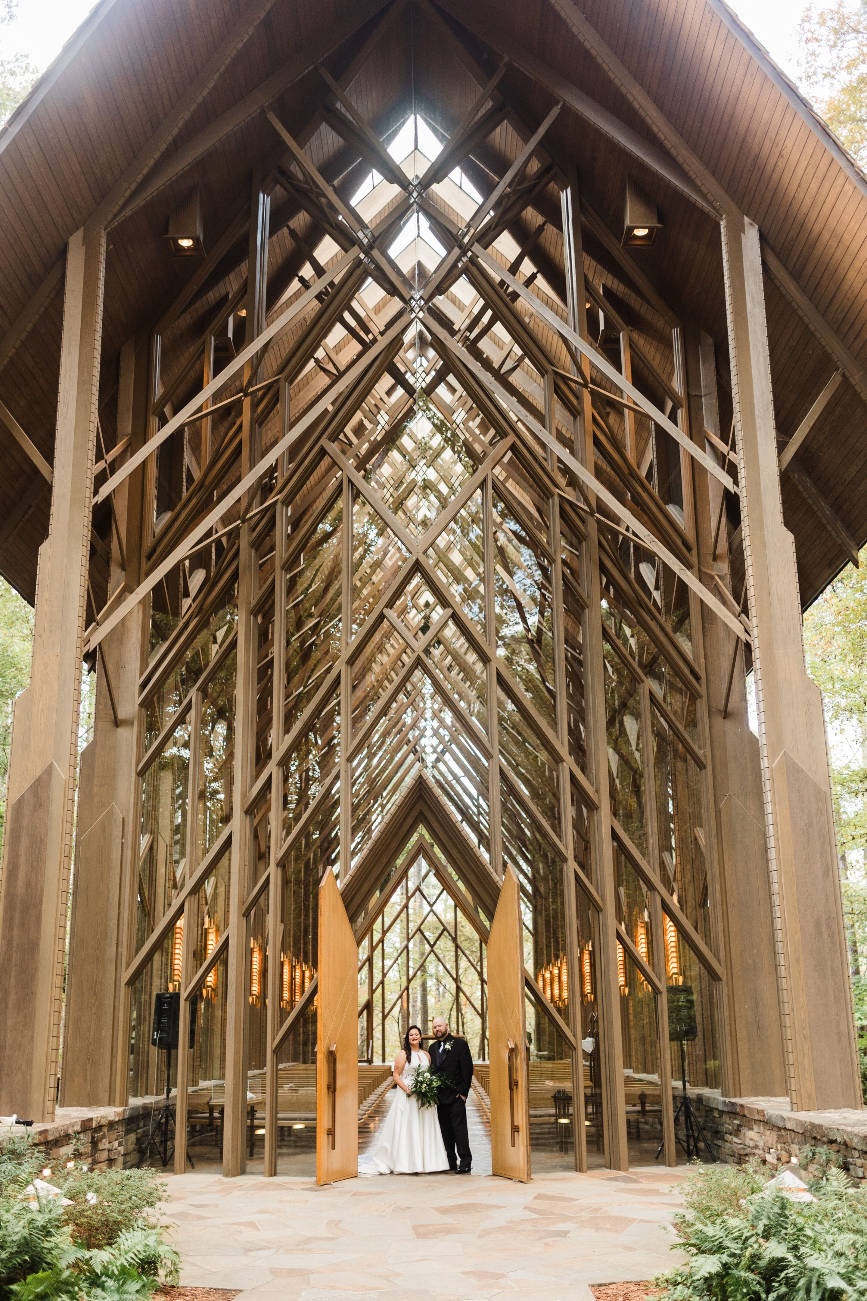 Angel & Trahern A bride and groom stand at the open doors of a modern glass chapel with wooden beams, surrounded by trees and natural light streaming in through the tall windows.