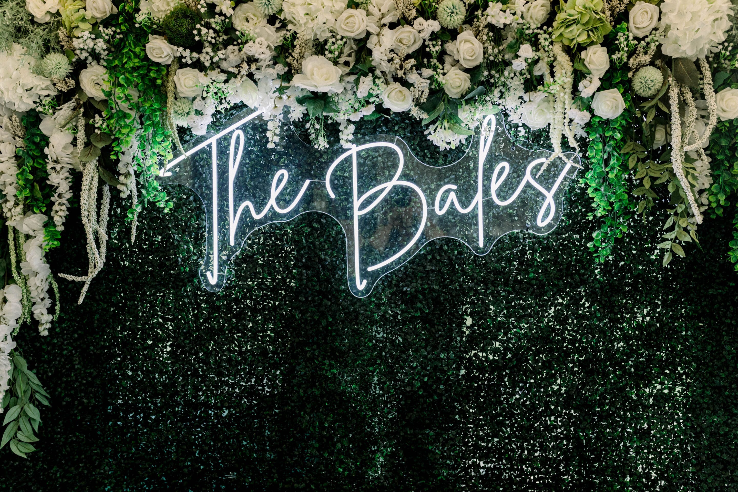 Keely + Joshua A neon sign reading The Bales is displayed on a lush green faux hedge wall, adorned with white roses, greenery, and floral decorations at the top.