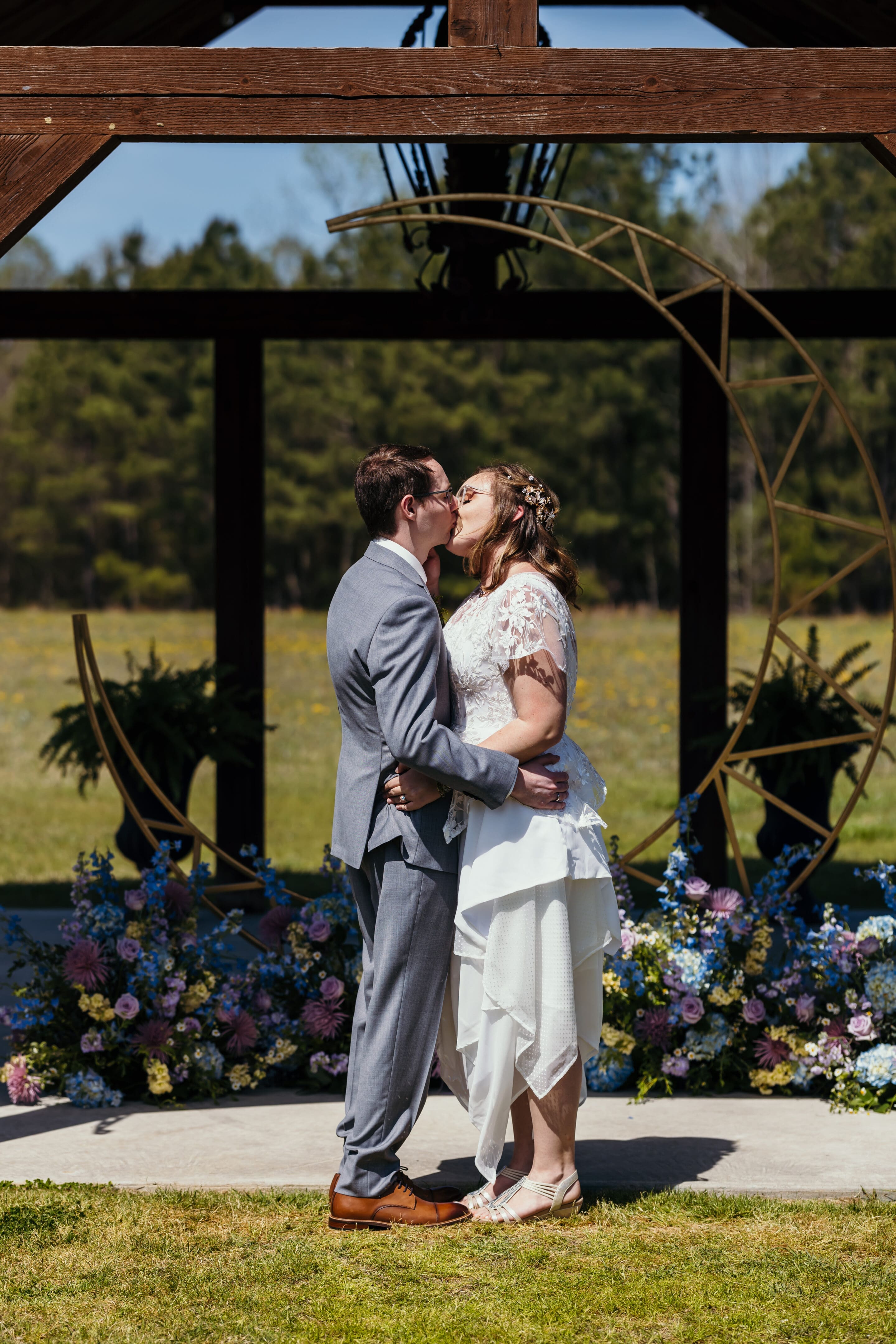 4 - Moon Themed Wedding A bride and groom kiss under a wooden arch decorated with flowers and a gold circular frame, surrounded by colorful floral arrangements outdoors on a sunny day.