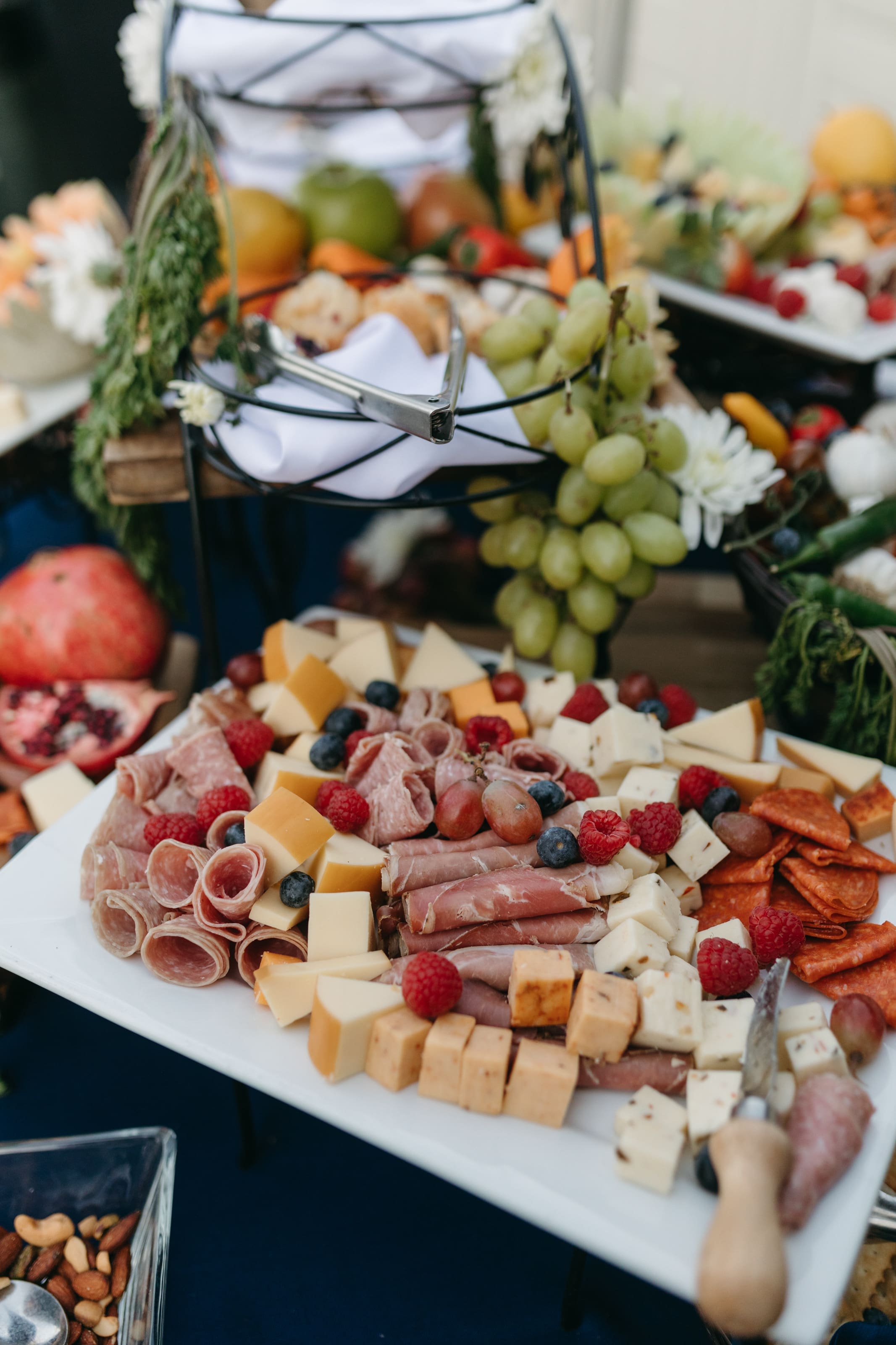 John David + Lindsey A platter with assorted cheeses, cured meats, berries, and grapes sits on a table, surrounded by pomegranate halves, nuts, and more fruit, creating an inviting charcuterie spread.