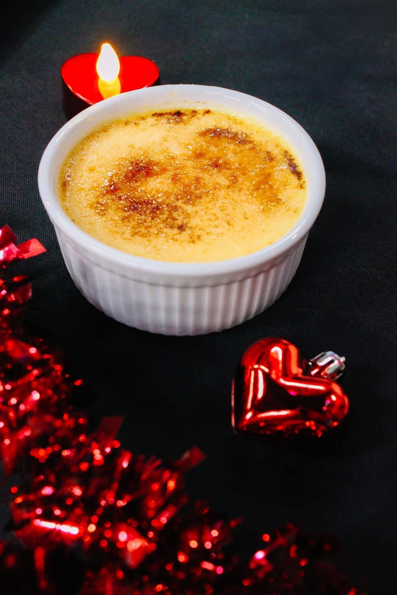 4 - Wedding Cake Crème Brûlée Recipe A ramekin of crème brûlée on a dark surface, with a lit red candle, a shiny red heart ornament, and red tinsel decorations nearby.