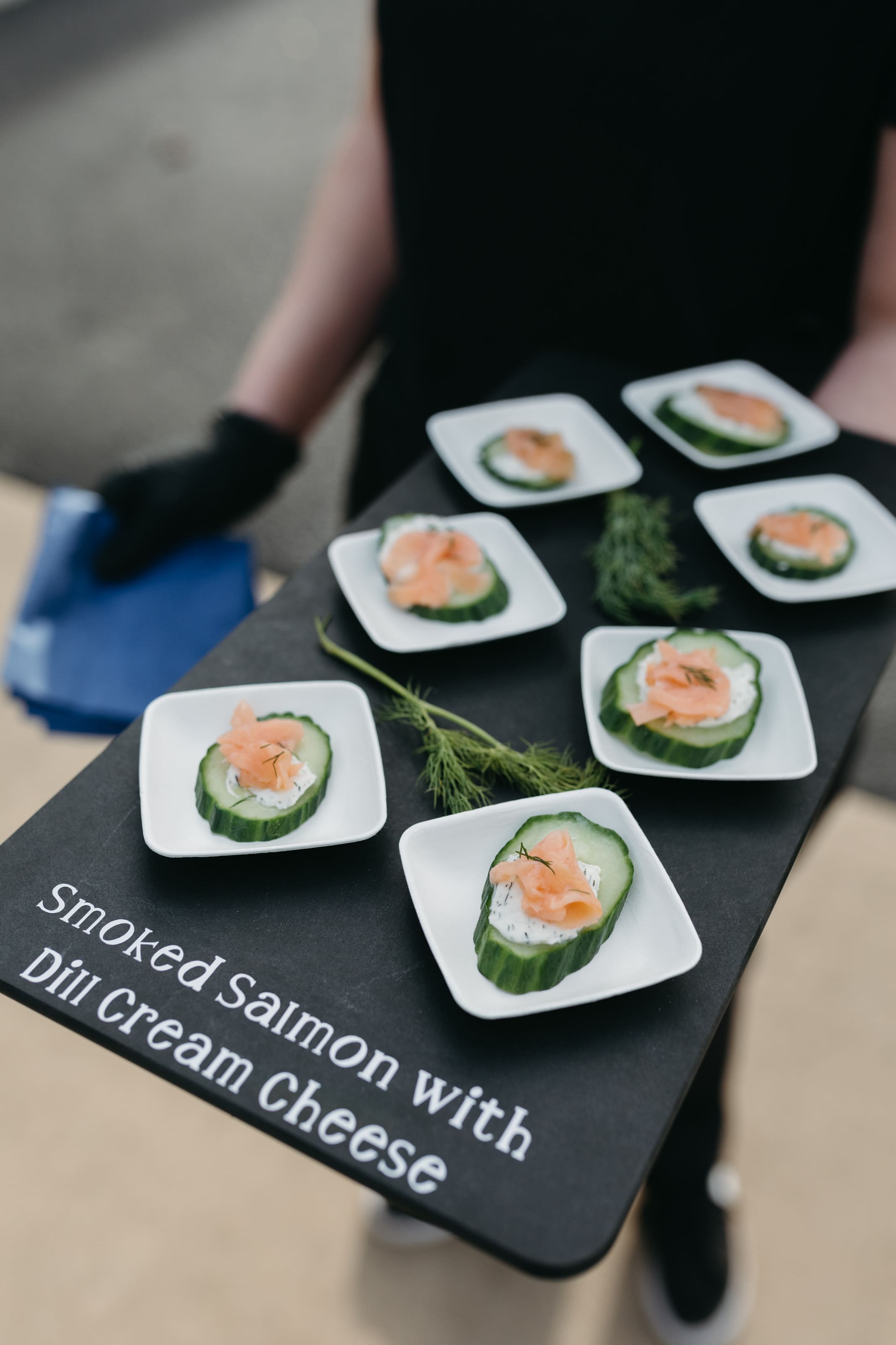 John David + Lindsey A server holds a tray with small plates of smoked salmon and dill cream cheese on cucumber slices, garnished with fresh dill. The tray text reads, Smoked Salmon with Dill Cream Cheese.