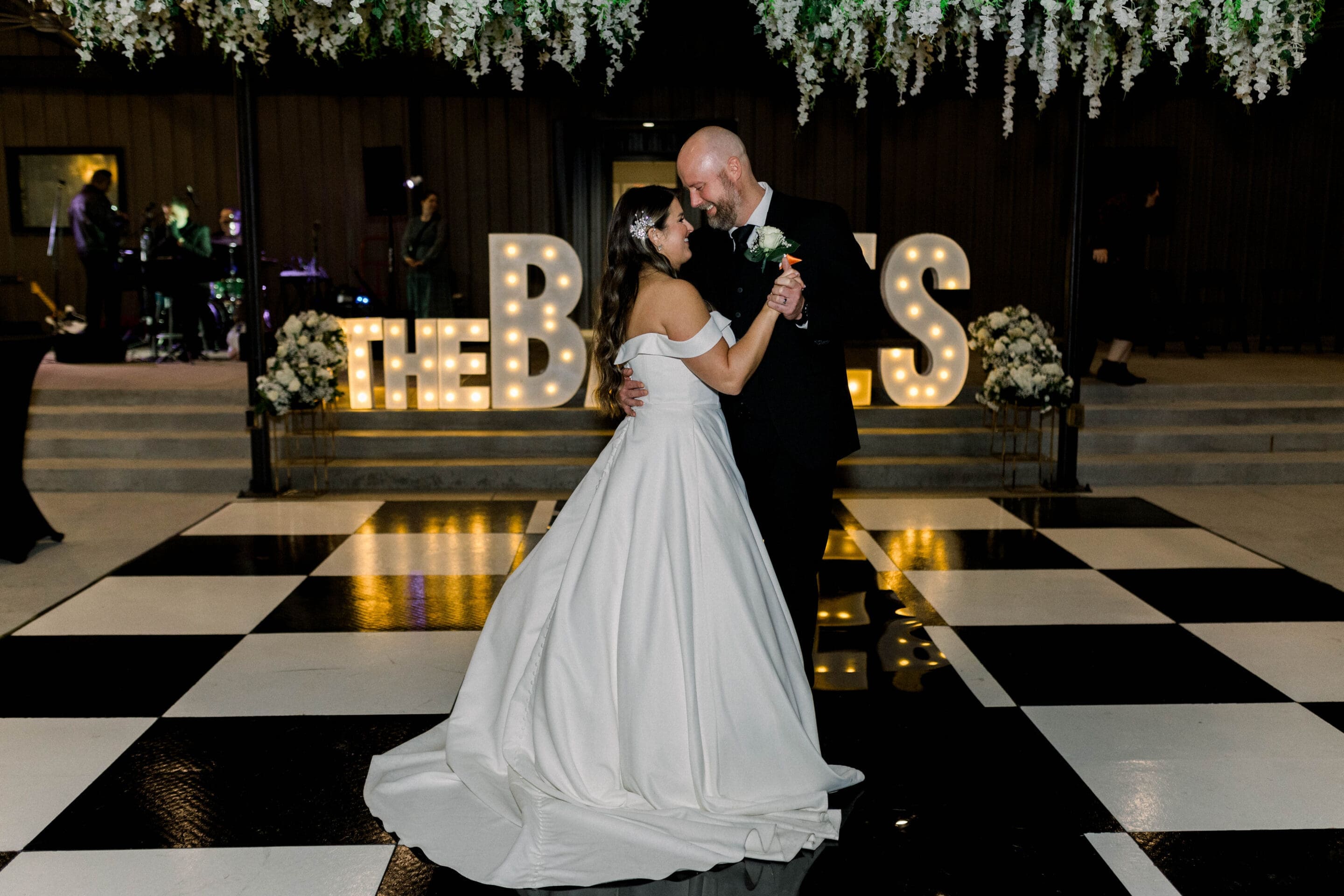 Keely + Joshua A bride and groom share a first dance on a black-and-white checkered floor, with floral arrangements and illuminated THE BOSS letters in the background. White flowers hang overhead, and a band performs off to the side.