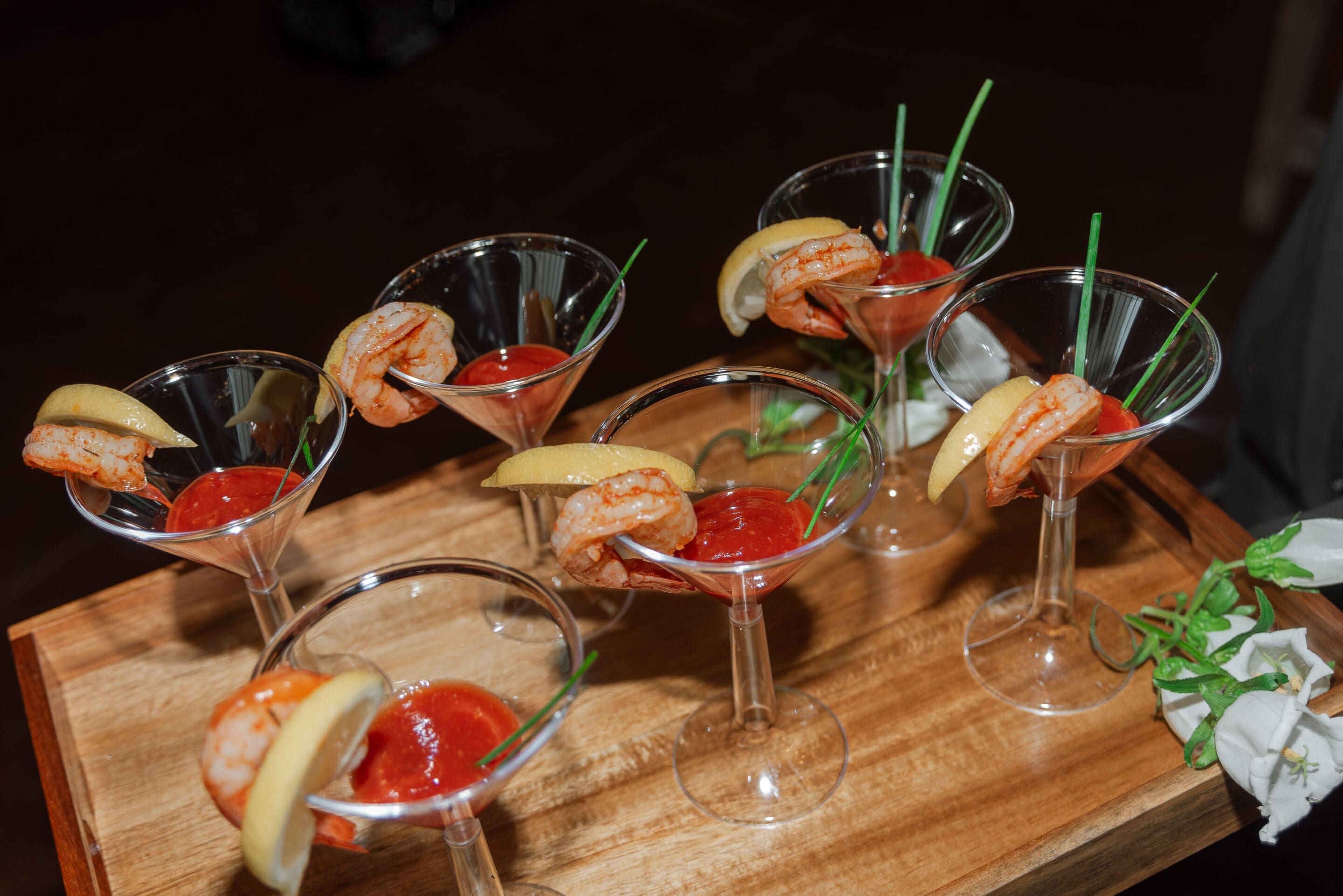 Lauren & Christian Six martini glasses filled with cocktail sauce, each garnished with a shrimp, a lemon wedge, and a piece of chive, are arranged on a wooden tray. White flowers are visible at the corner of the tray.
