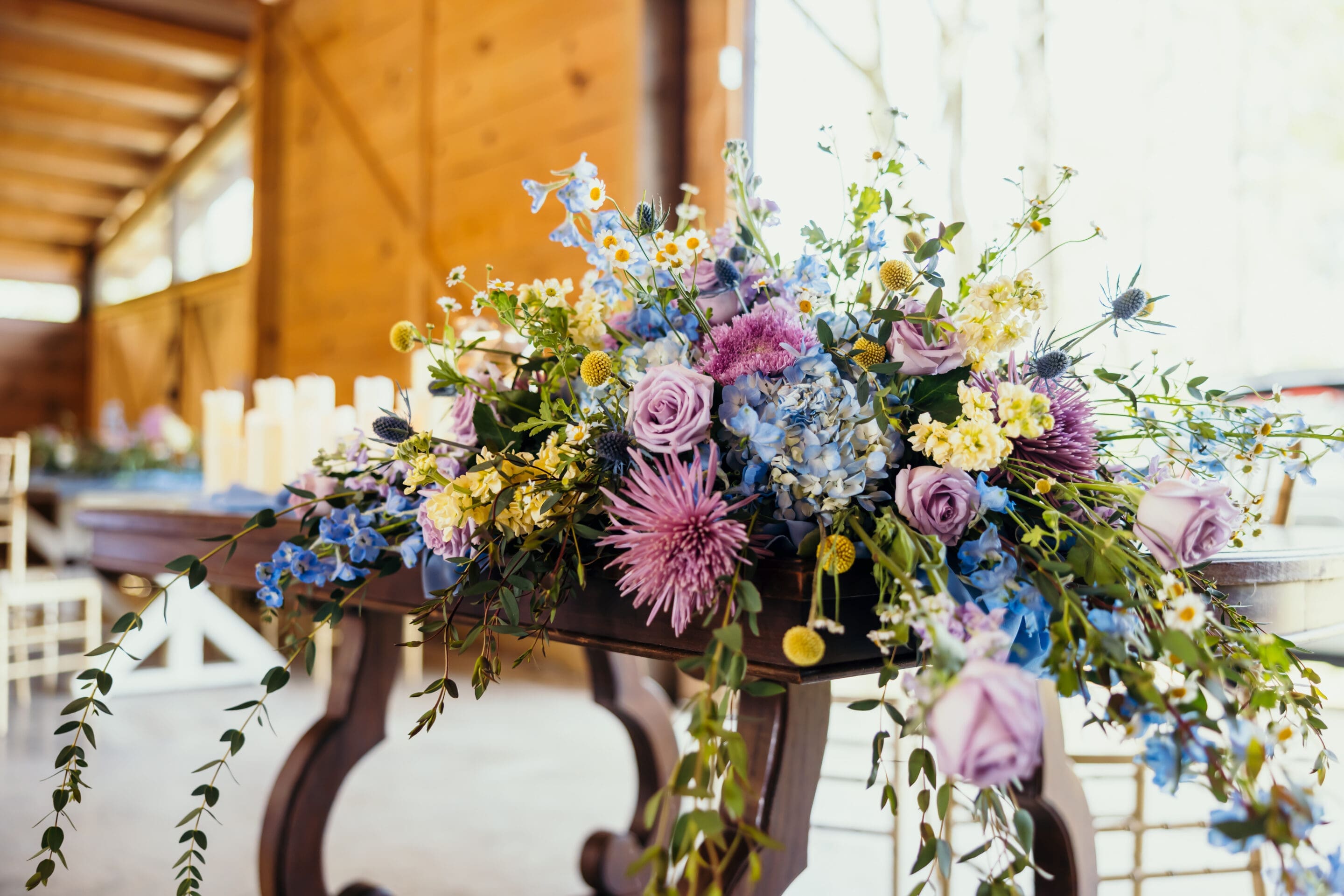 5 - Sky Themed Wedding Florals A wooden table decorated with a lush floral arrangement, featuring purple roses, blue hydrangeas, yellow and white flowers, and greenery, set in a warmly lit rustic room with wooden walls.