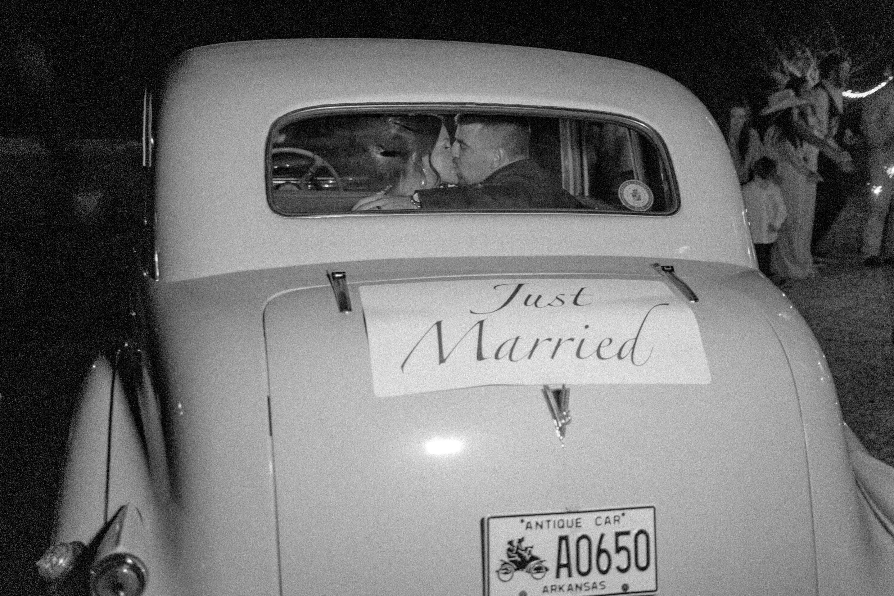 Antique Car Getaway Car Little Rock A black and white photo of a couple in a vintage car at night, seen from behind. A Just Married sign is taped to the back, and the car has an Arkansas antique license plate. People are visible in the background.
