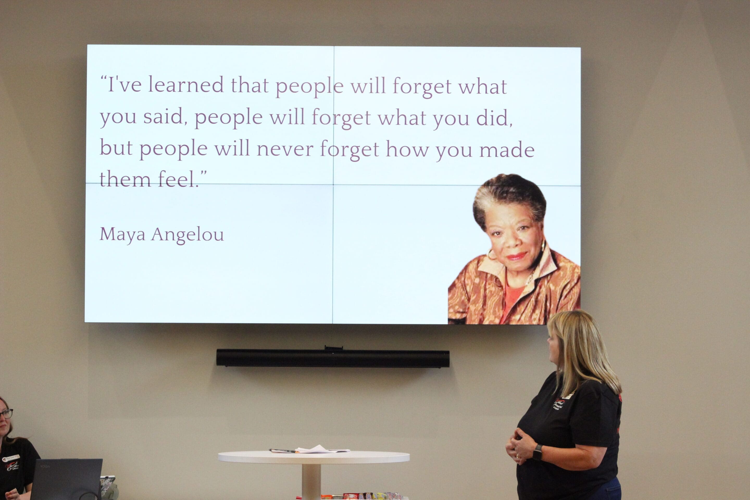 How to Create a Positive Workplace A woman stands in front of a large screen displaying a quote by Maya Angelou and her photo. The quote reads, Ive learned that people will forget what you said, people will forget what you did, but people will never forget how you made them feel.