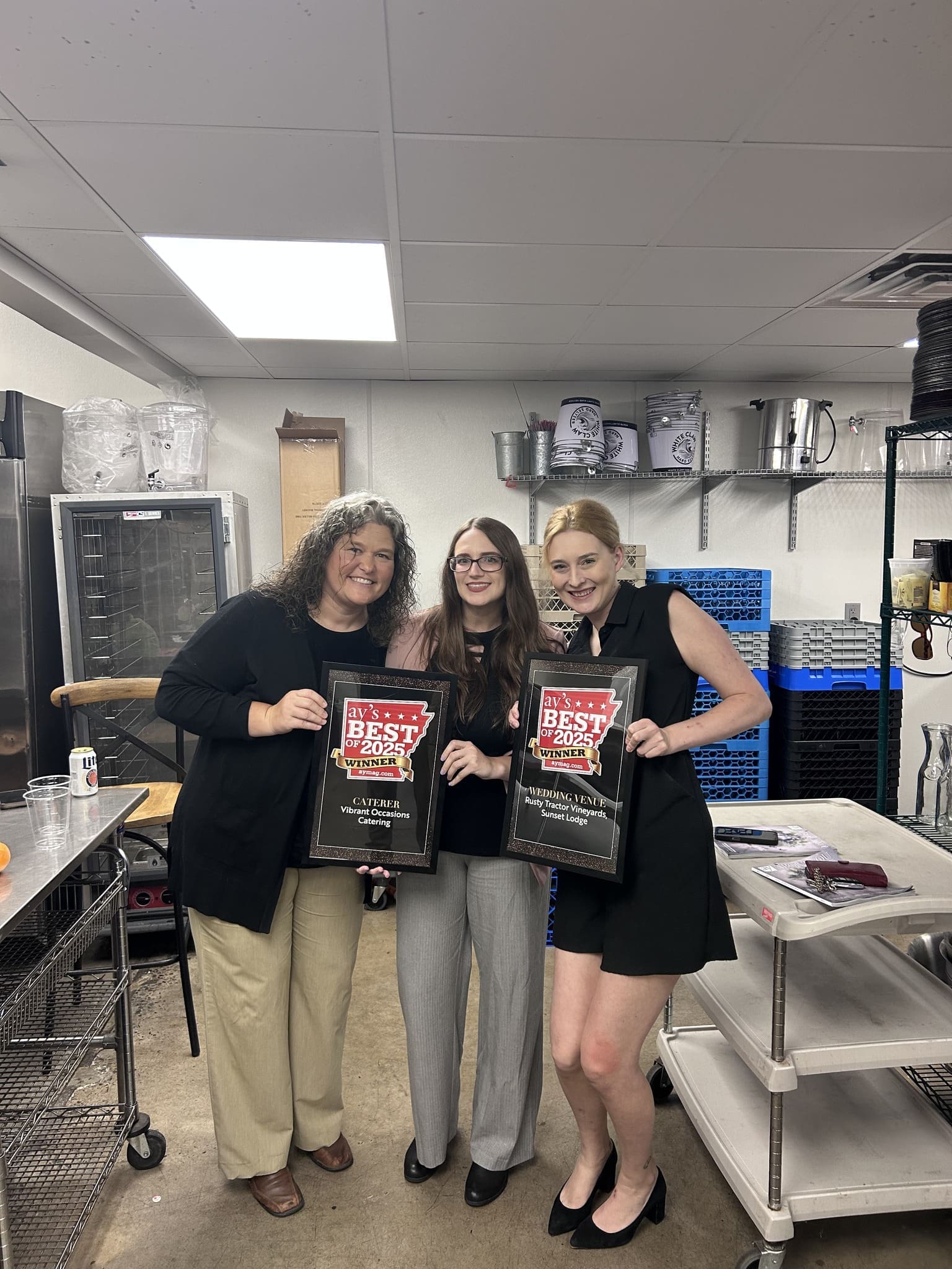 AY Magazine's Best Of 2025 Party at Rusty Tractor Vineyards Three women standing in a commercial kitchen, smiling and holding framed Best of the Best awards. Kitchen equipment and shelves are visible in the background.