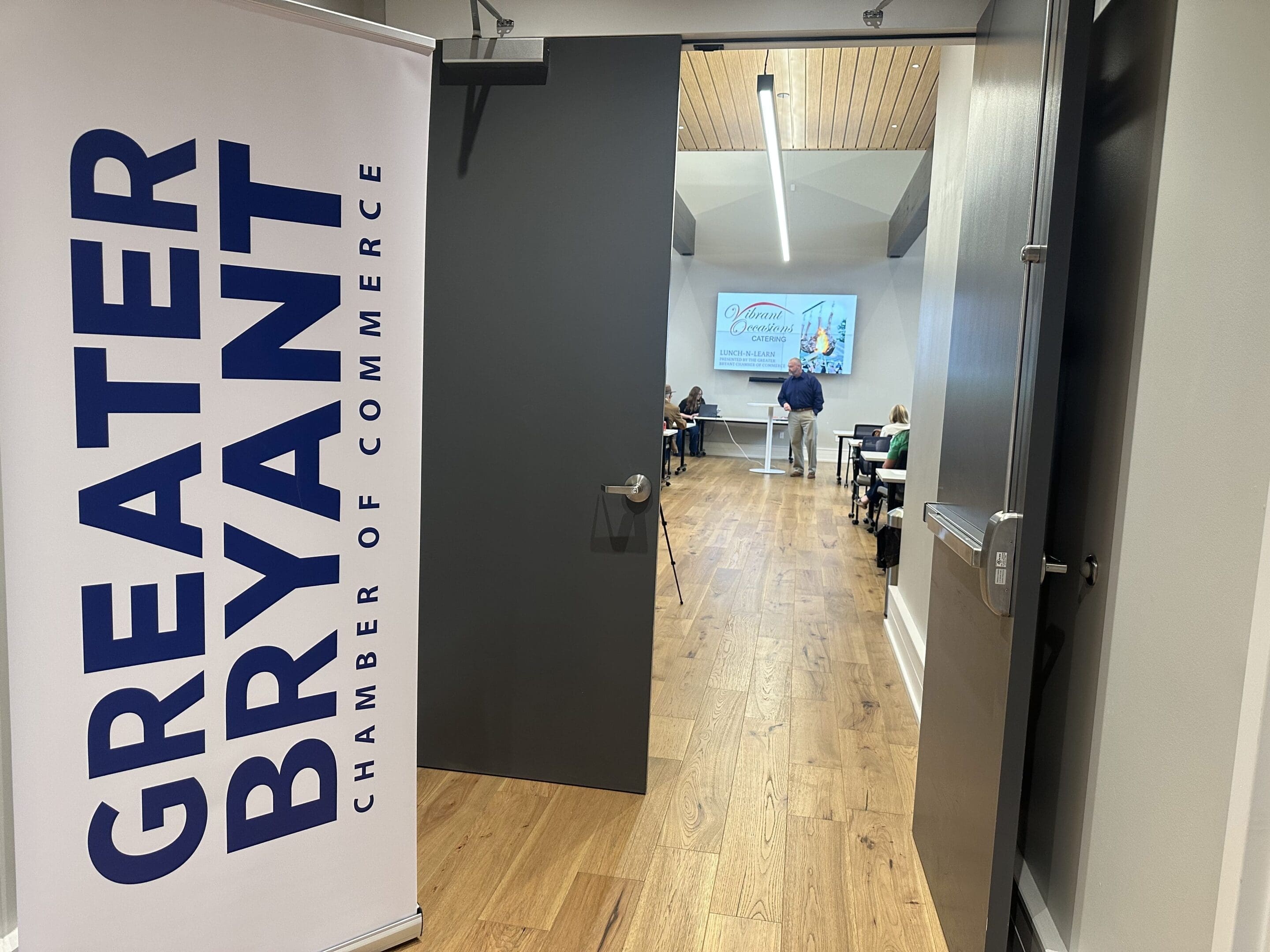 Bryant Chamber of Commerce Heart Leadership A door is open to a meeting room with people seated at tables facing a presenter and a projected screen. A sign by the door reads Greater Bryant Chamber of Commerce. The room has wood flooring and modern decor.