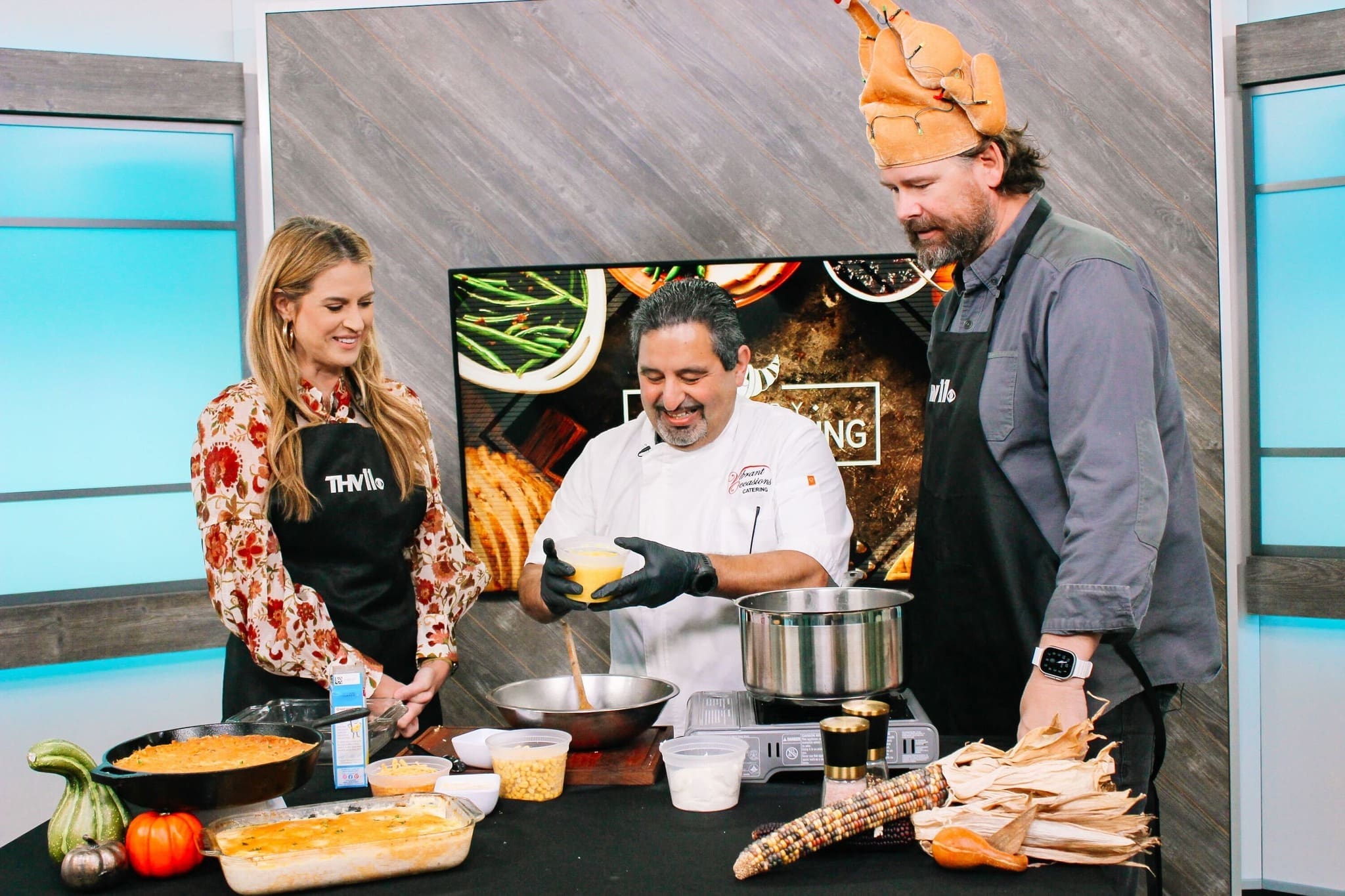 Celebrity Chef THV11 Little Rock Three people stand in a kitchen studio cooking together. The chef in the center prepares food while smiling, as the man on the right wears a turkey hat. The woman on the left and the man both wear black aprons. Various foods are on the counter.
