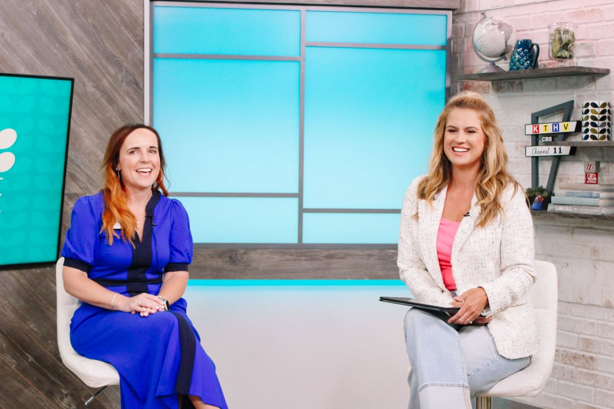 Two women sit and smile in a modern studio setting; one wears a blue dress, the other a white blazer and pink top, holding a tablet. The background features bright blue panels, shelves, and a TV screen.