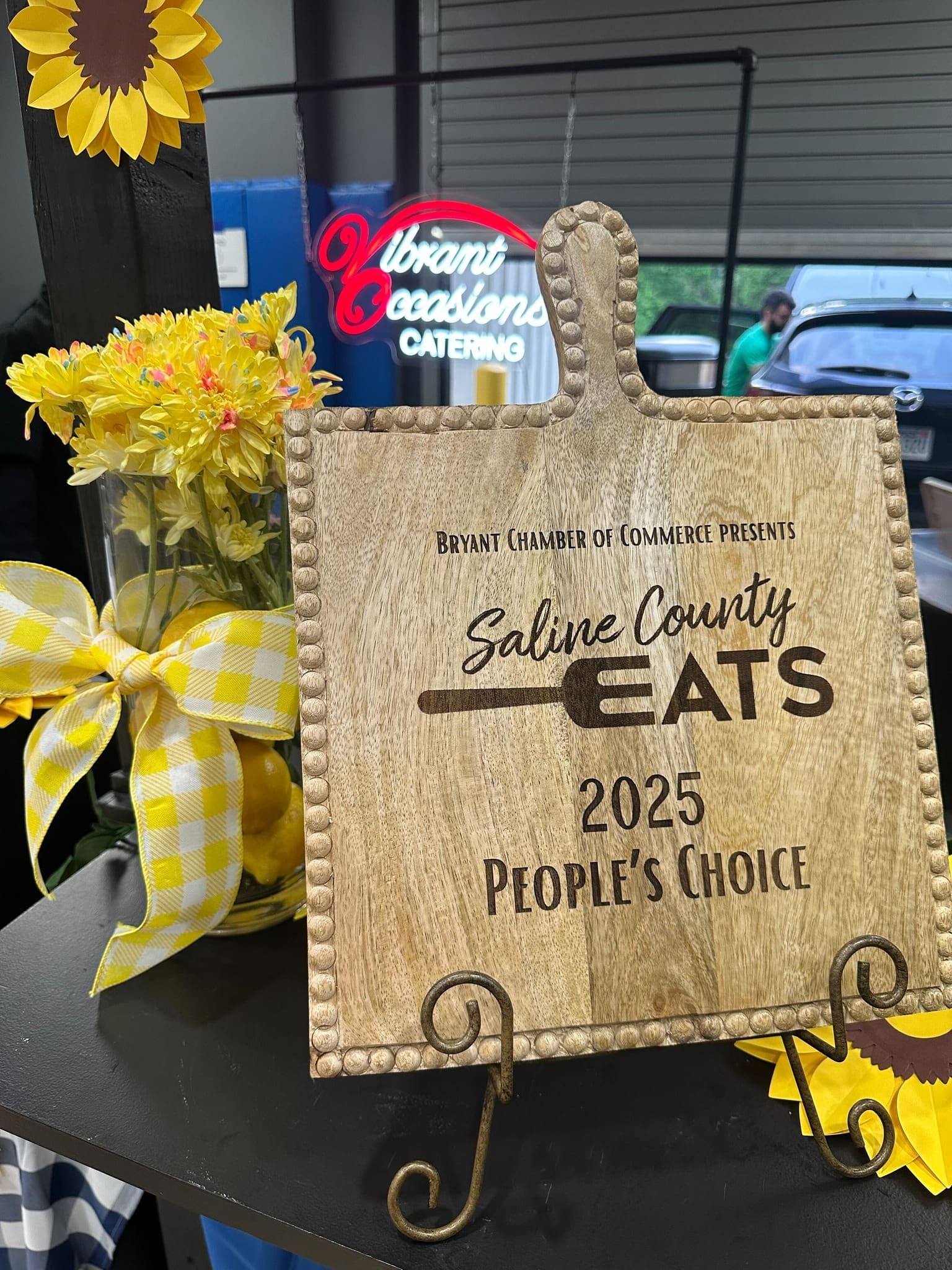 Saline County Eats 2025 People_s Choice Winner A wooden plaque reads “Saline County Eats 2025 People’s Choice” from the Bryant Chamber of Commerce. It’s displayed on a stand next to a mason jar of yellow flowers with a yellow plaid bow, and a sunflower decoration.