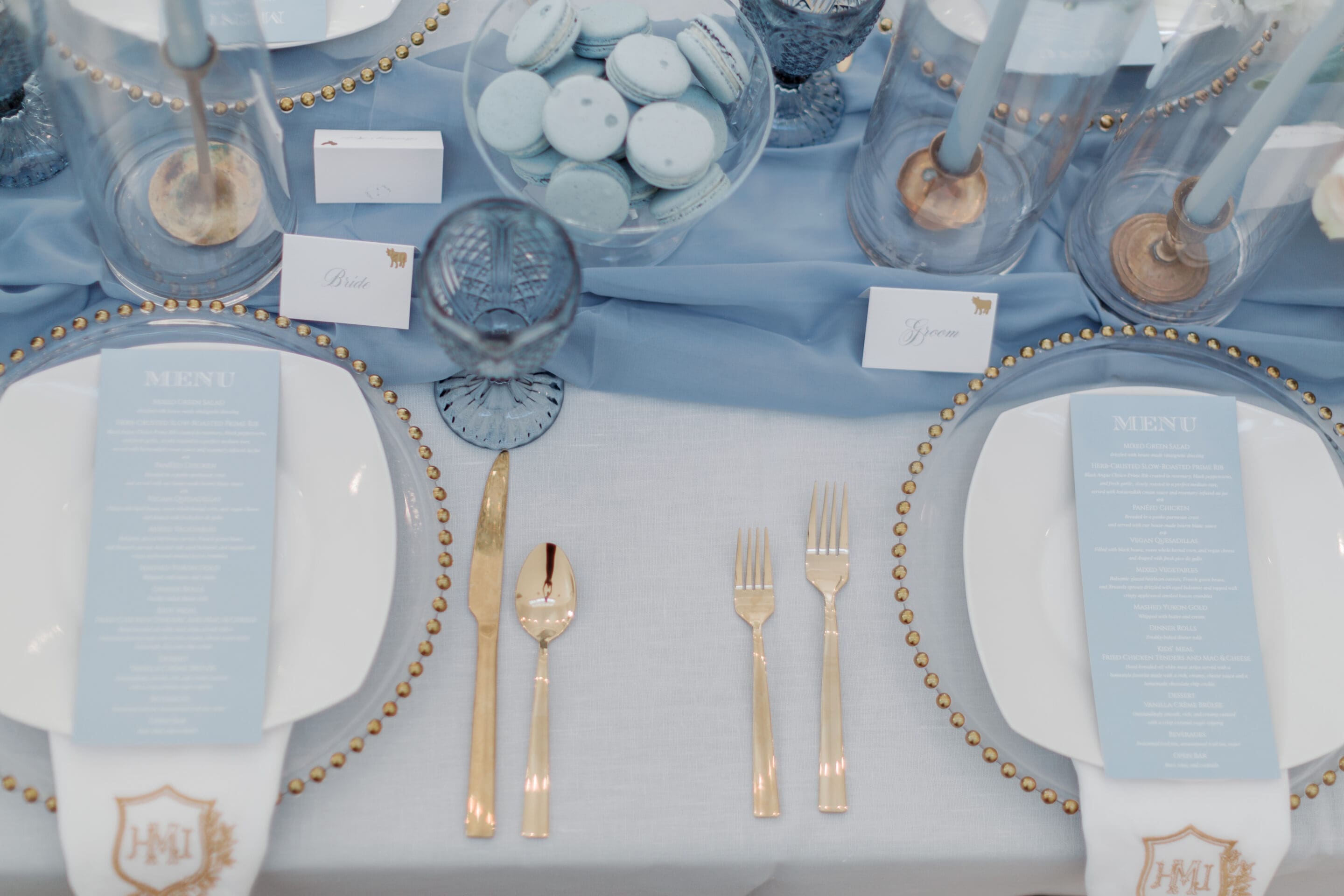 Best Wedding Caterer in Little Rock Elegant table setting with gold cutlery, white plates, blue menus, blue glassware, and blue macarons on a centerpiece. Blue and white cards mark places at a table with a pale blue tablecloth and blue candles.