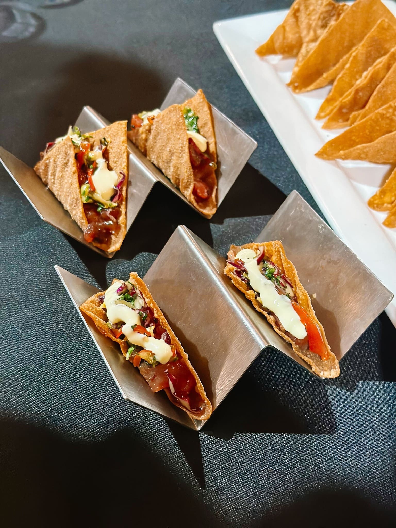 Three mini tacos filled with diced vegetables, herbs, and a drizzle of creamy sauce are displayed in metal taco holders on a black surface; tortilla chips are on a white tray in the background.