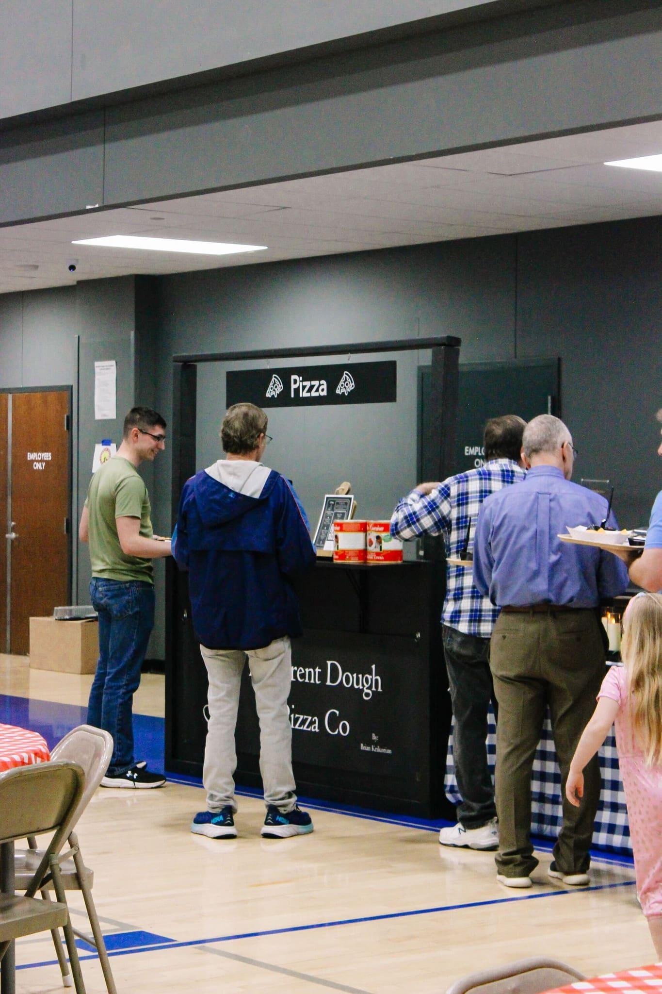 Different Dough Pizza Co. Saline County People stand in line at a pizza stall inside a gymnasium, waiting to be served. The stall has a sign that says “Pizza.” Nearby tables are covered with blue checkered cloths. A young girl stands in the foreground.