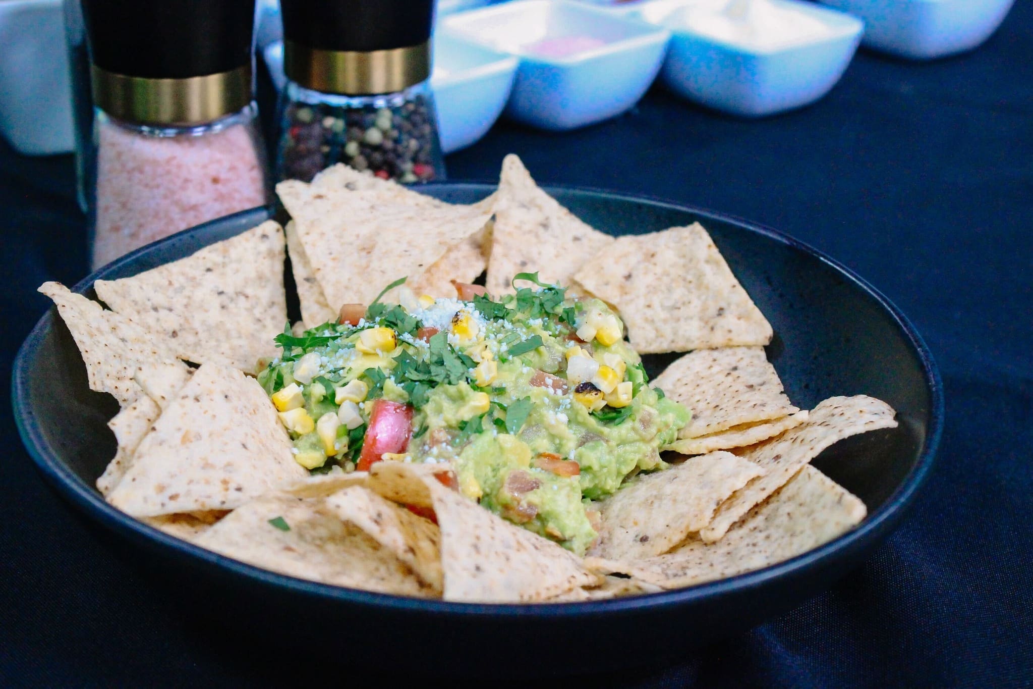A black bowl filled with tortilla chips and guacamole garnished with corn, tomatoes, onions, and cilantro—perfect for a fun guacamole bar. Seasonings and small condiment bowls are visible in the background.