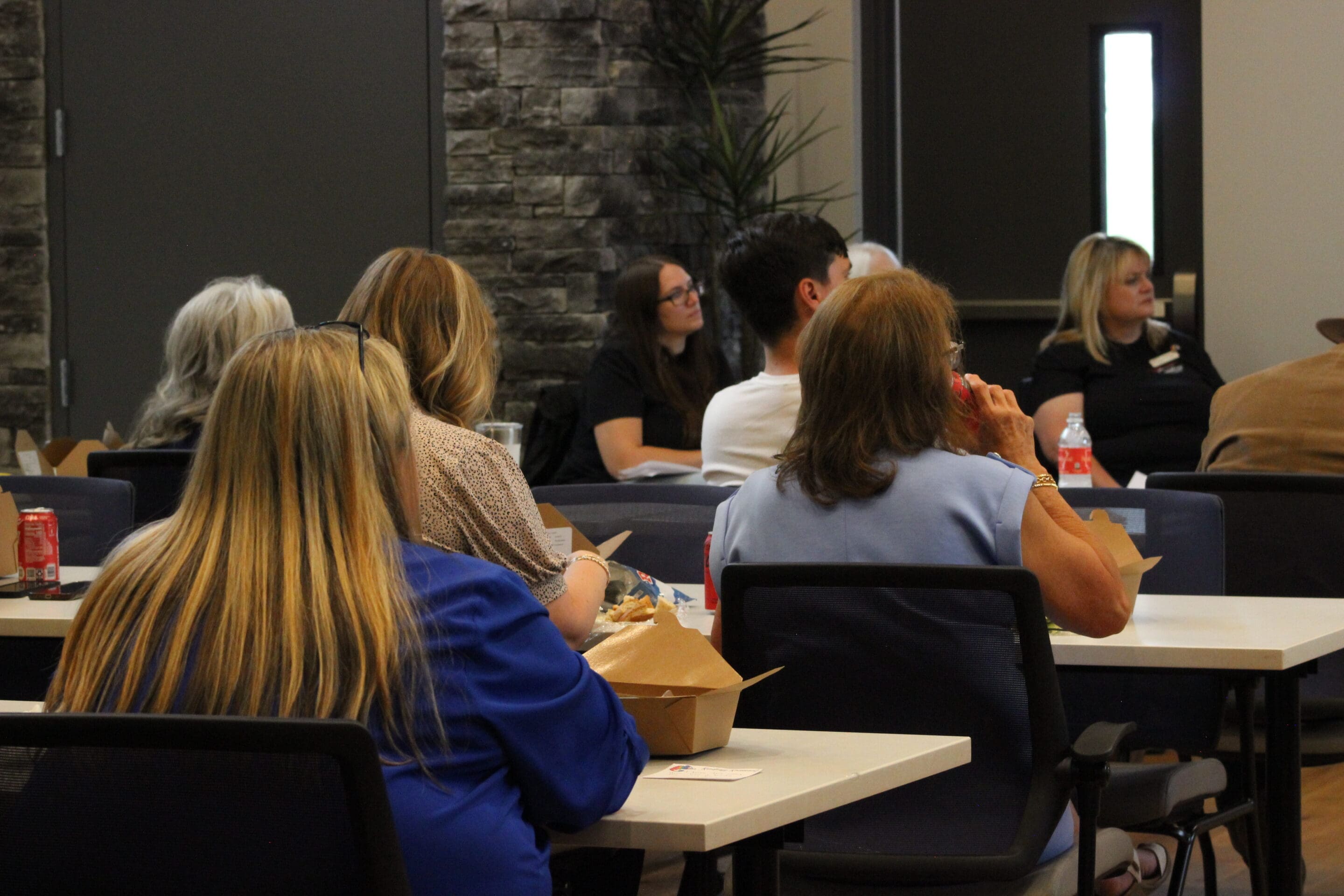 4 - Bryant AR Chamber MeetingJPG People sit at tables in a conference room, listening attentively. Some are eating from takeout boxes, with drinks like soda on the tables. The room has a modern design, with stone walls and plants visible.