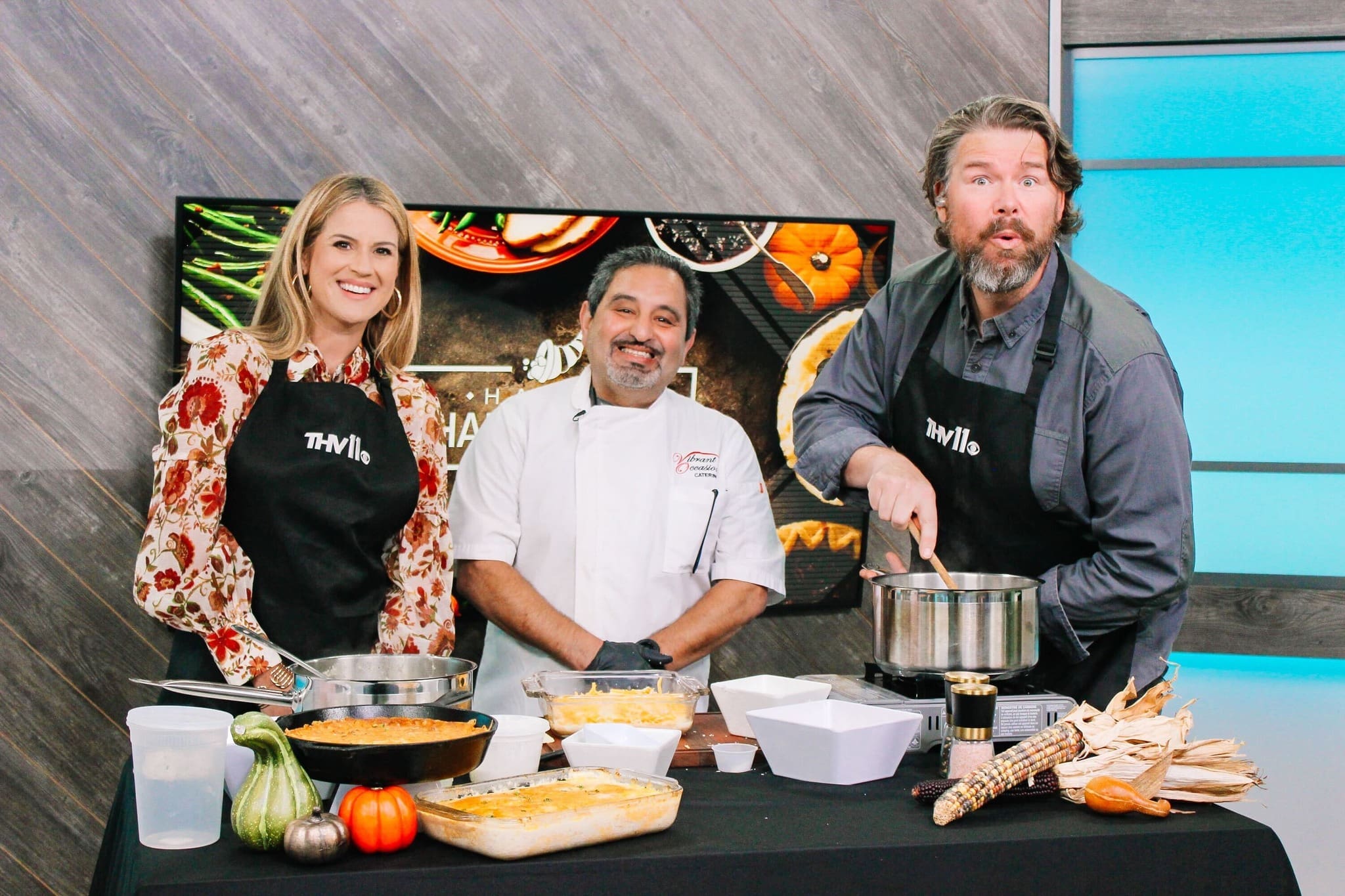 Chef Serge Krikorian Southern Food Three people stand behind a table with prepared dishes and cooking utensils. One stirs a pot, another stands smiling in a chefs coat, and the third smiles at the camera. A TV screen and fall decor are in the background.