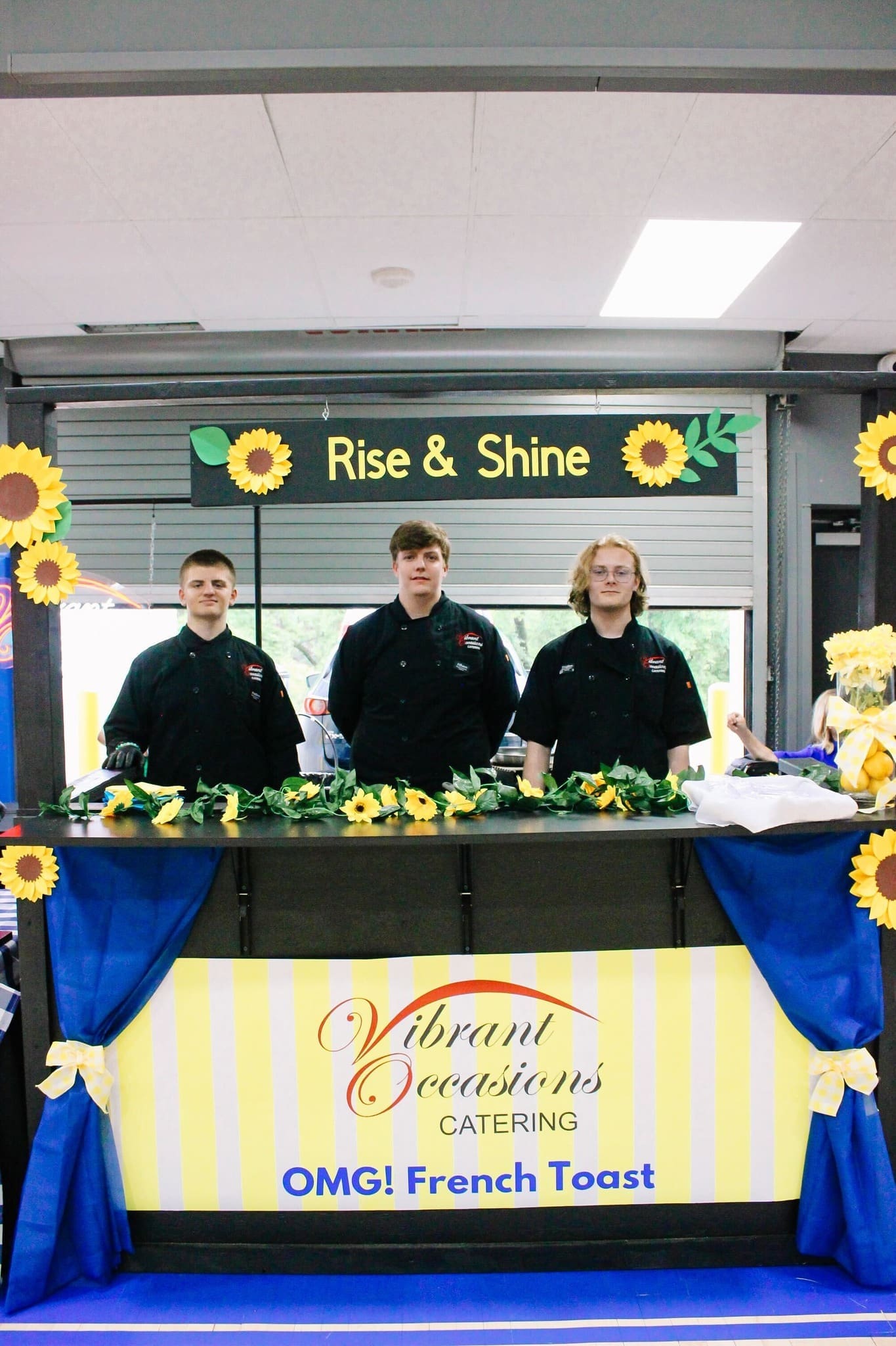 Formerly Called Taste of Bryant Three people in black uniforms stand behind a decorated booth with sunflowers and a sign reading “Rise & Shine.” The counter displays “Vibrant Occasions Catering: OMG! French Toast” on a yellow and white striped banner.
