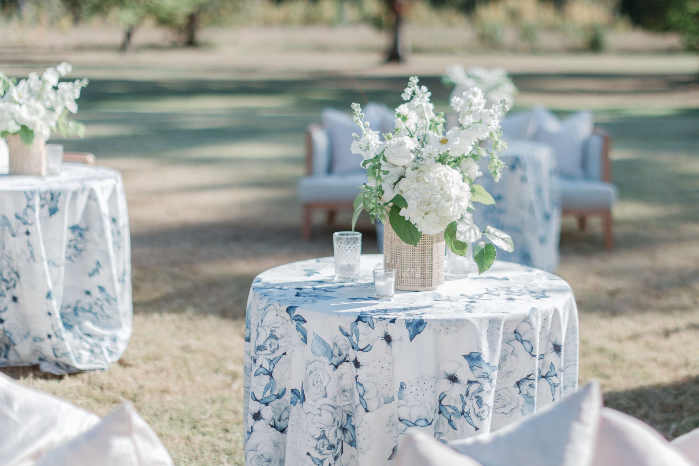 Cocktail Hour Ideas A round table with a blue and white floral tablecloth holds a wicker vase of white flowers and a glass candle holder, set outdoors on grass with armchairs and more decorated tables in the background.