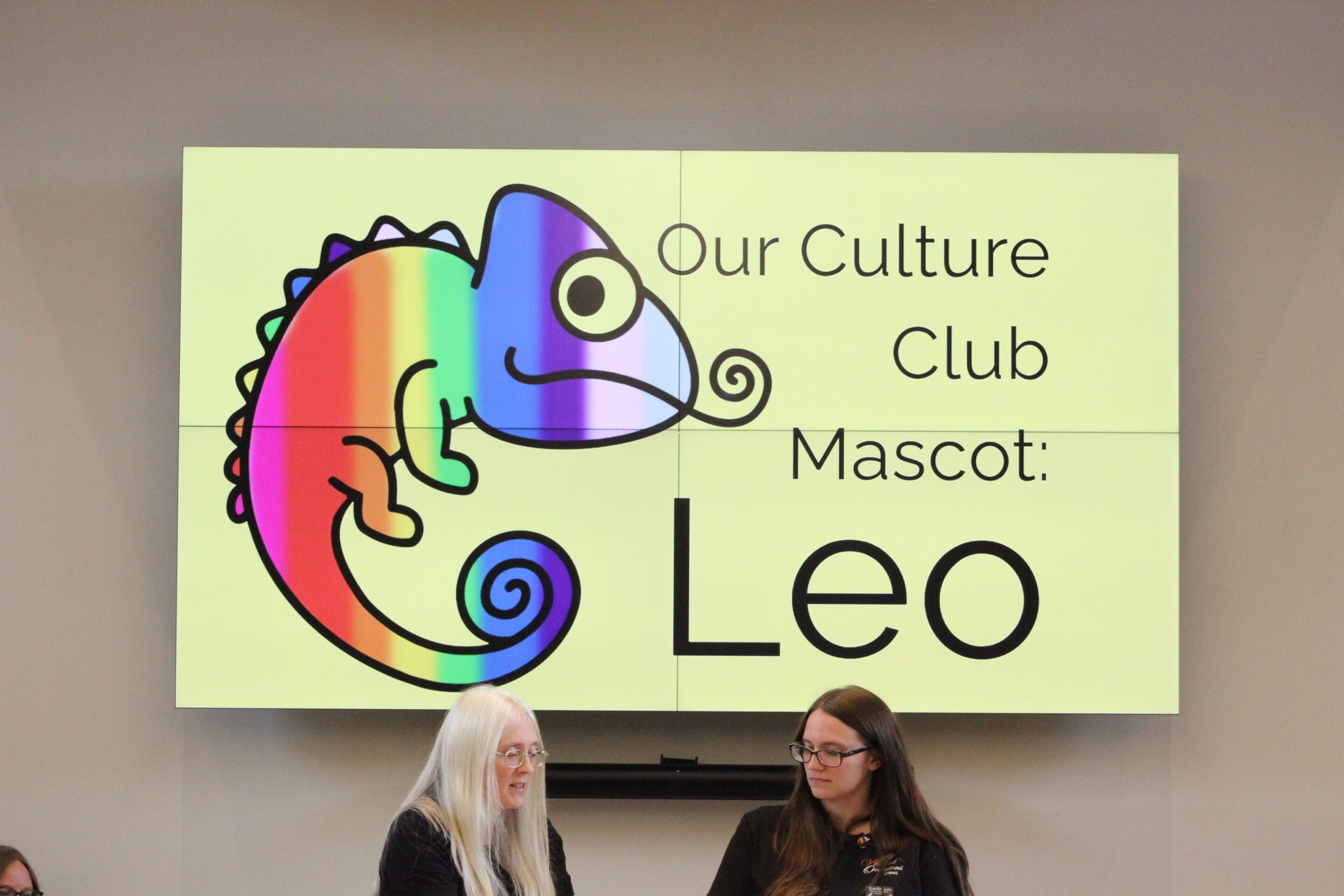 5 - Company Culture Leaders A large screen displays a colorful chameleon with the text Our Culture Club Mascot: Leo. Two women with long hair sit in front of the screen, one with white hair and one with brown hair.