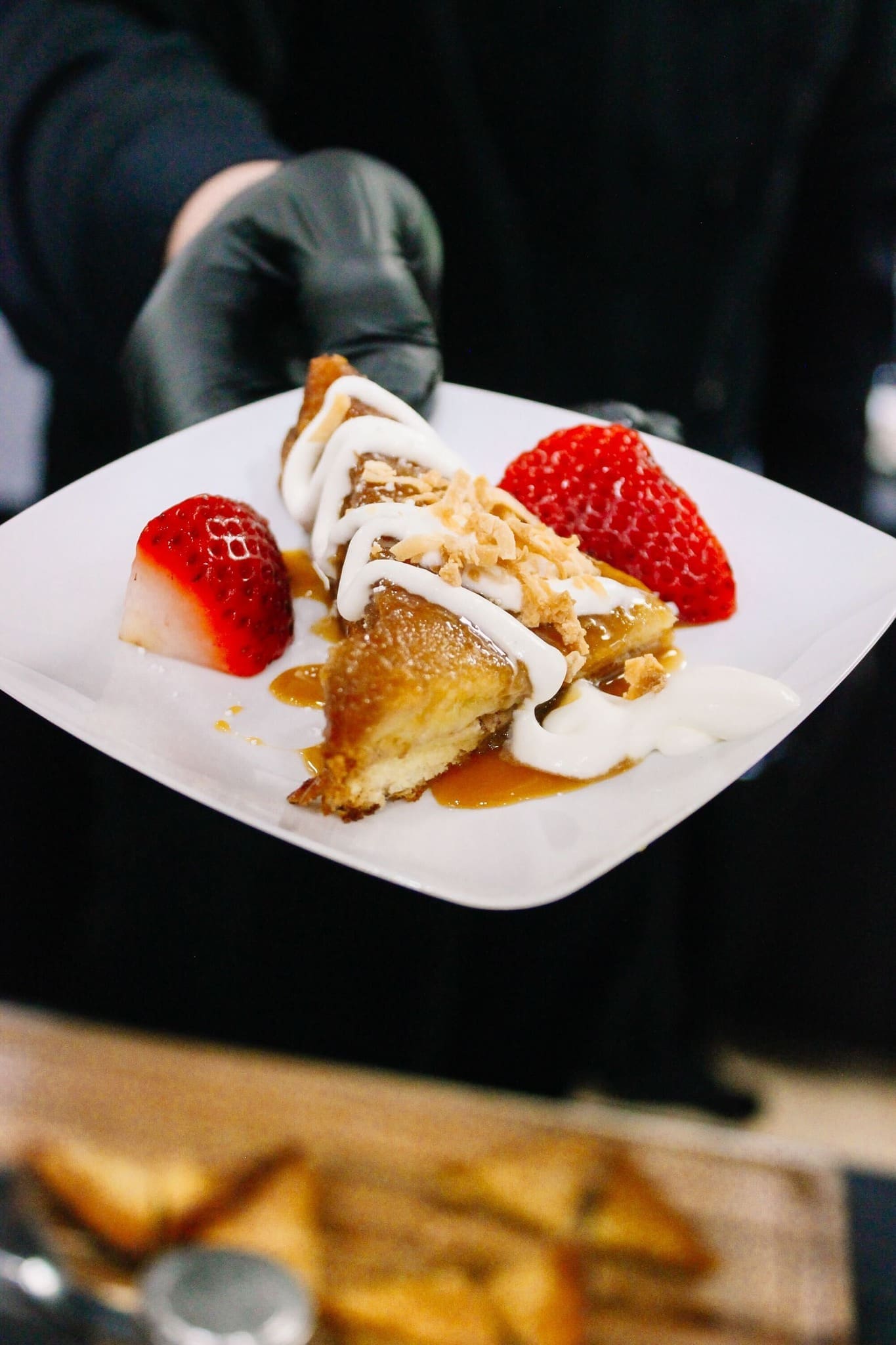 5 - OMG French Toast by Vibrant Occasions Catering A person wearing a black glove holds a plate with a slice of dessert topped with white icing, caramel sauce, chopped nuts, and served with two strawberry halves.