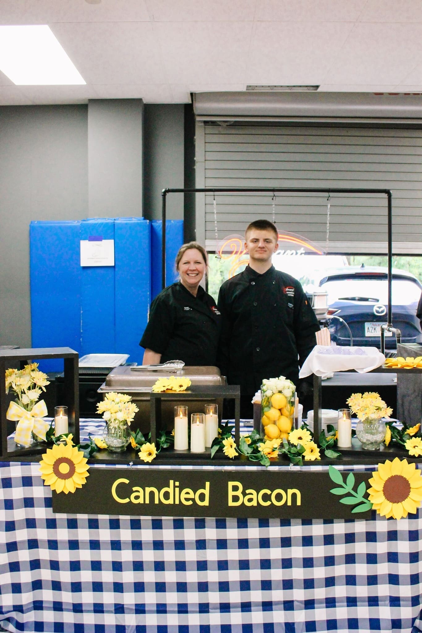 Best Saline County Caterer Two people in black chef coats stand behind a “Candied Bacon” booth decorated with sunflowers, lemons, candles, and a blue-checkered tablecloth. Plates of food are set out on the table.