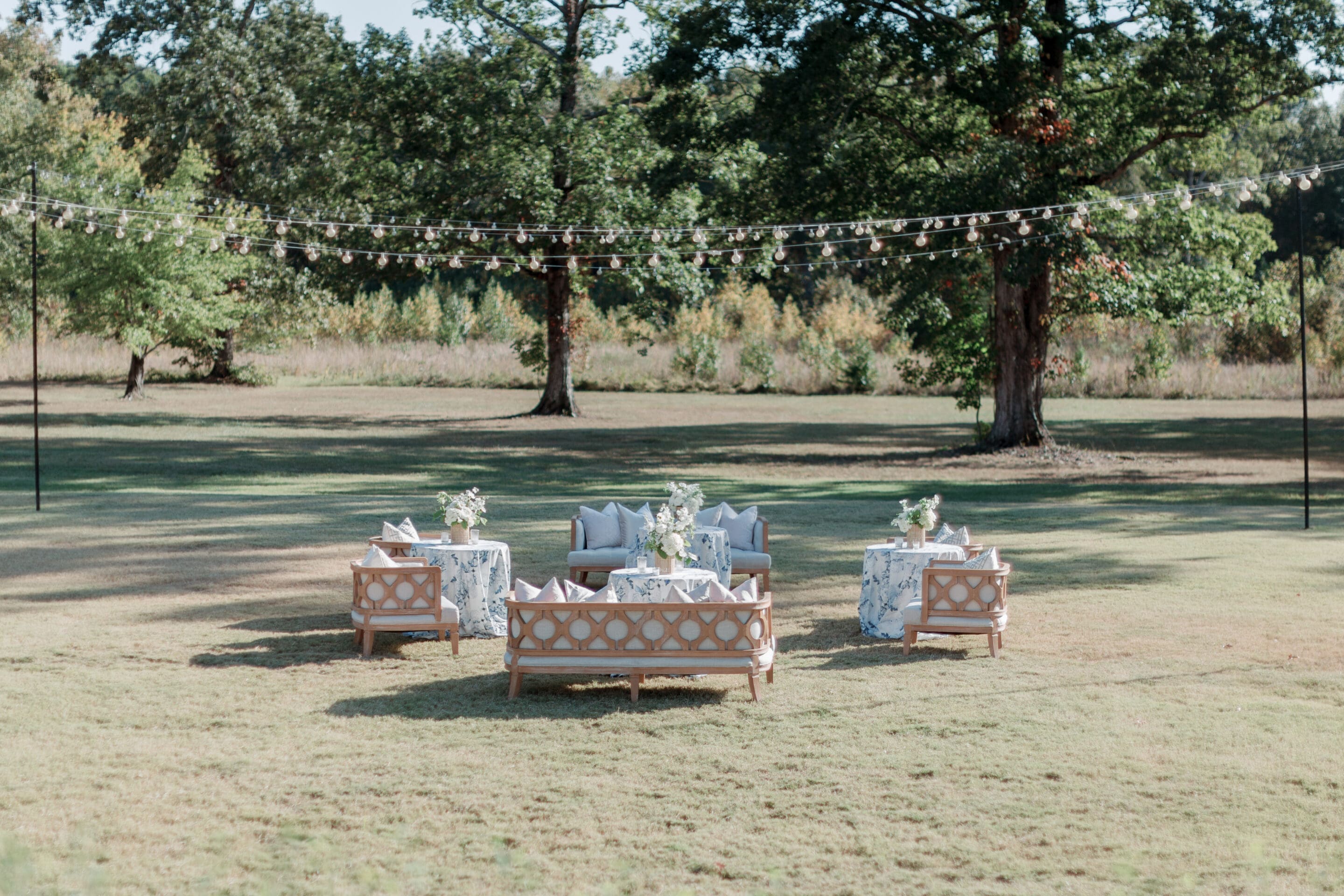How to Set Up Cocktail Hour Outside Four sofas with patterned blue and white tablecloth-covered tables form a lounge area on a grassy lawn, surrounded by trees and string lights, perfect for an Elegant Spring Wedding at The Grandeur House in Little Rock.