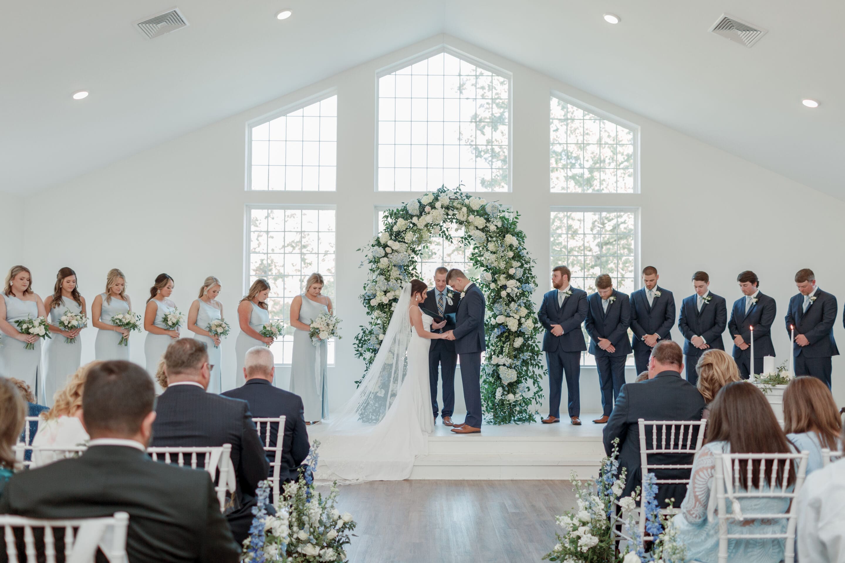 Delia Jeffcoat Wedding Planner A bride and groom stand at the altar holding hands, surrounded by their wedding party in a bright, flower-adorned chapel with large windows and seated guests watching the ceremony.
