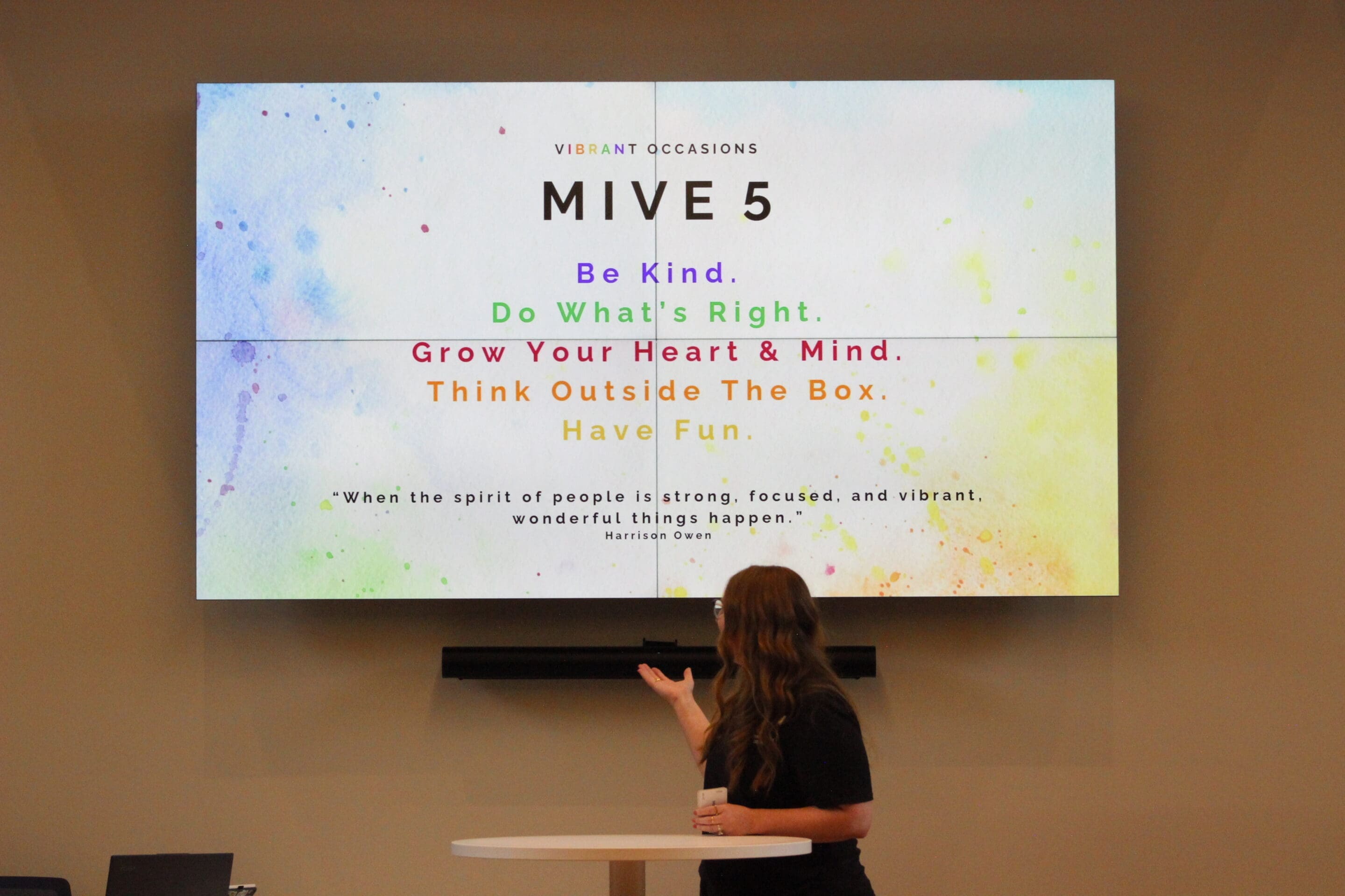 How to Write a Mission Statement A woman gestures toward a colorful presentation slide displaying MIVE 5 with the phrases: Be Kind, Do Whats Right, Grow Your Heart & Mind, Think Outside The Box, and Have Fun, along with a motivational quote.