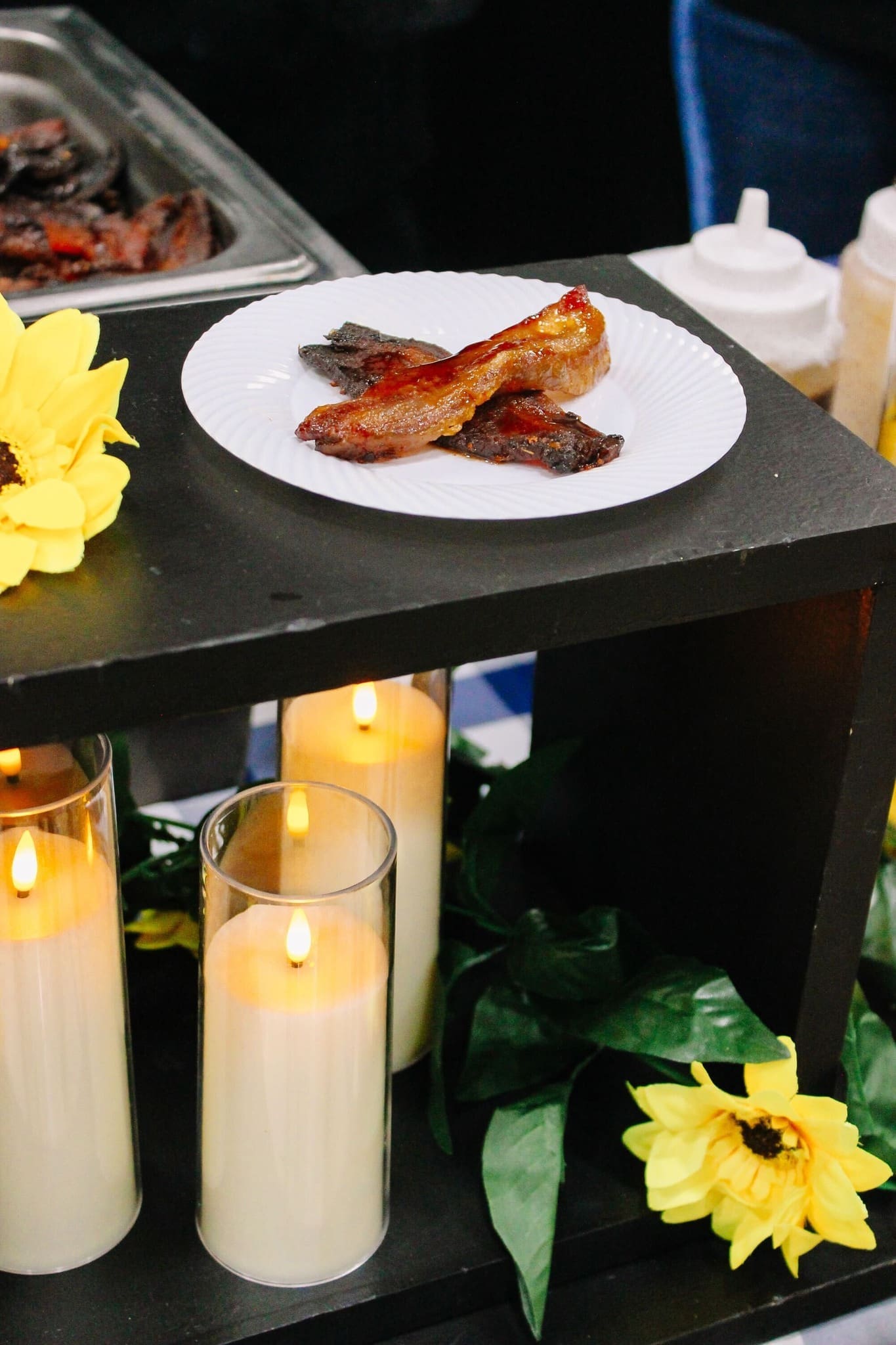 7 - Who is the Best Caterer in Central Arkansas A white plate with several pieces of cooked bacon sits on a black shelf decorated with artificial sunflowers, green leaves, and lit pillar candles in glass holders. Squeeze bottles are visible in the background.