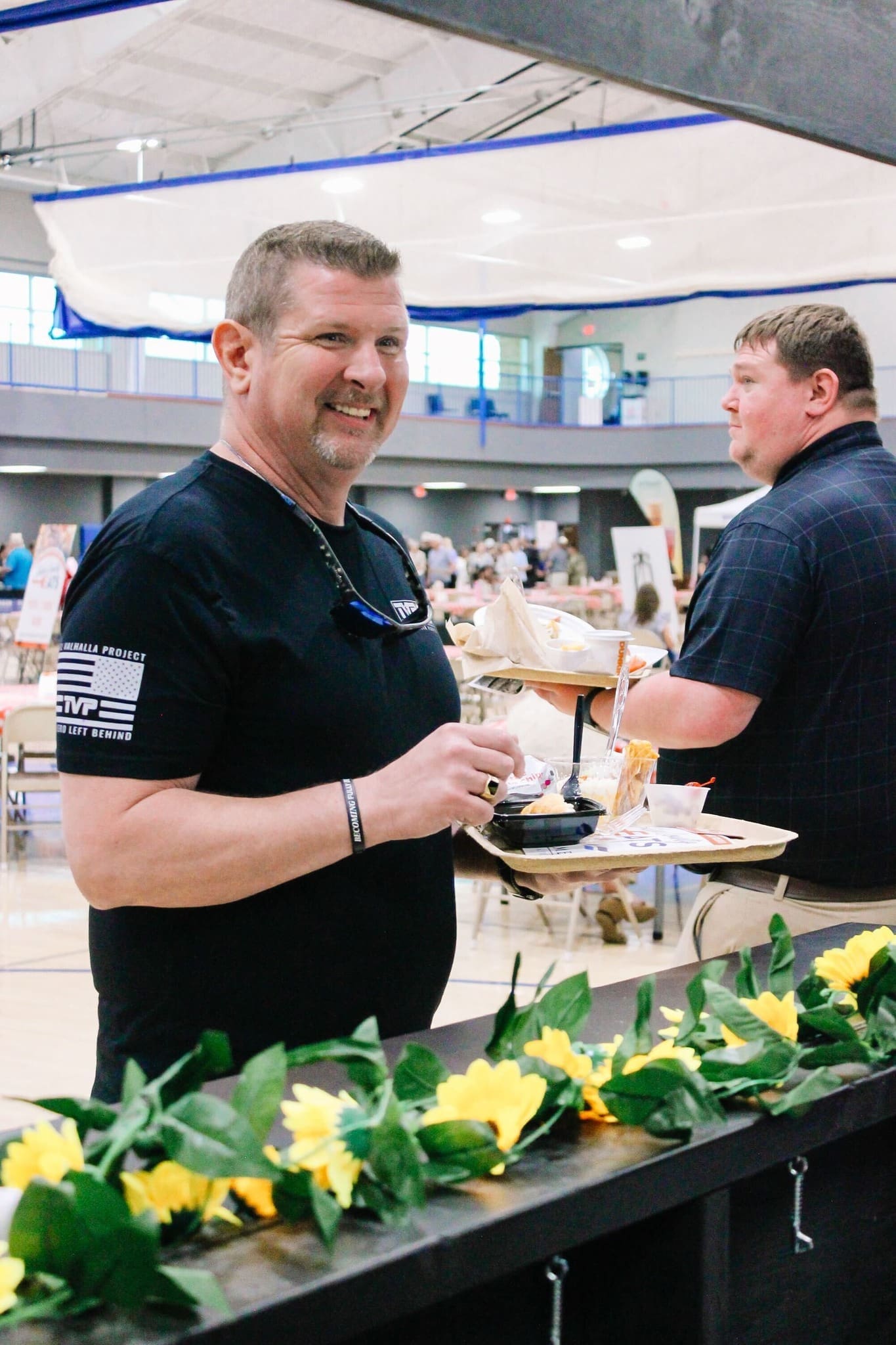 Who is the Best Caterer in Saline County A smiling man in a black t-shirt holds a plate of food at an indoor event. Another man stands behind him, and sunflowers decorate the foreground. The background shows tables and people in a gymnasium setting.