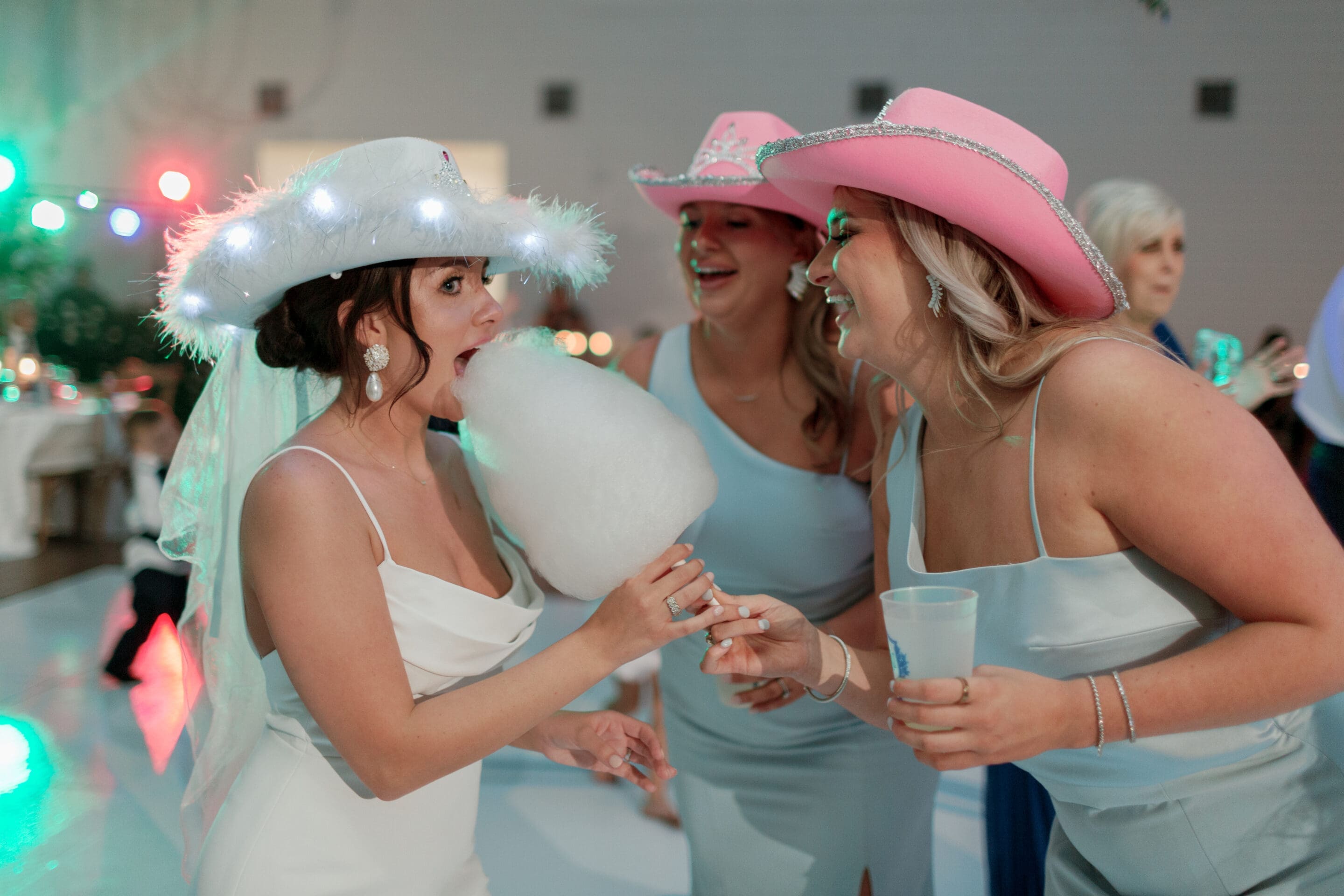 Wedding Ideas in Arkansas A bride in a white dress and light-up hat eats cotton candy while laughing with two bridesmaids in pink cowboy hats and light blue dresses at a lively indoor celebration.