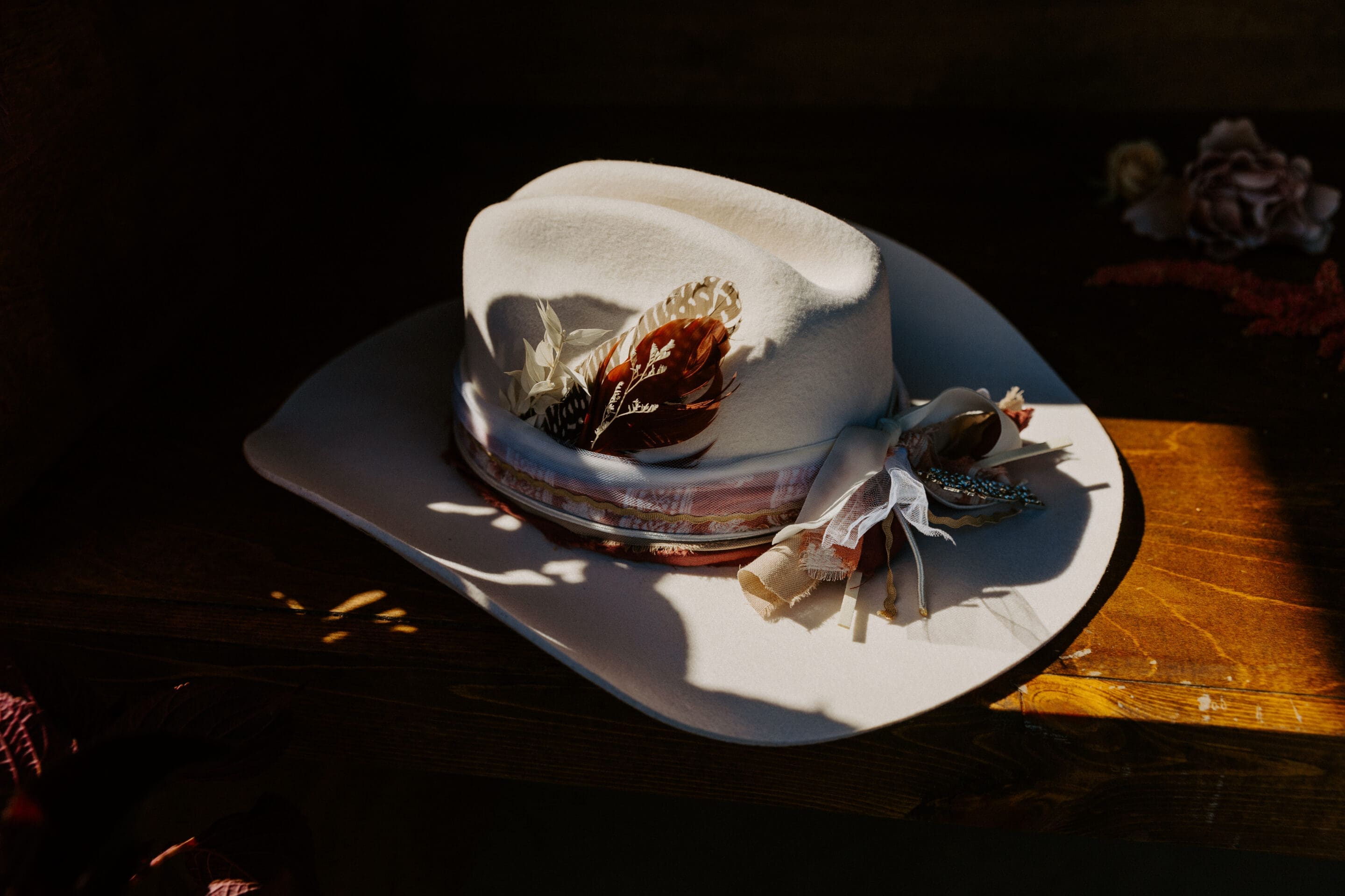 A white cowboy hat decorated with ribbons, lace, and a feather rests on a wooden surface in warm, natural light.
