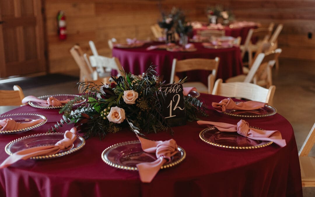 A round table with a burgundy tablecloth, set with clear plates, pink napkins, and a floral centerpiece featuring roses and greenery. A sign reads Table 12. Other similarly set tables are in the background.