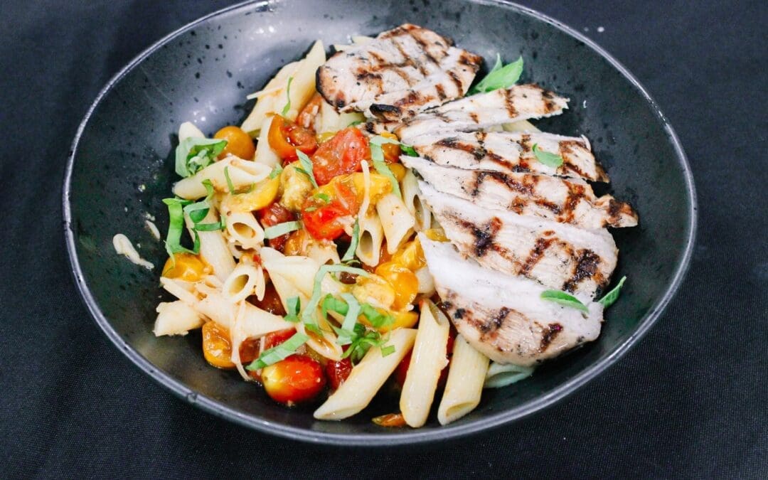 A black bowl containing penne pasta with colorful cherry tomatoes and fresh herbs, served alongside sliced grilled chicken breast with visible grill marks, set on a dark background.